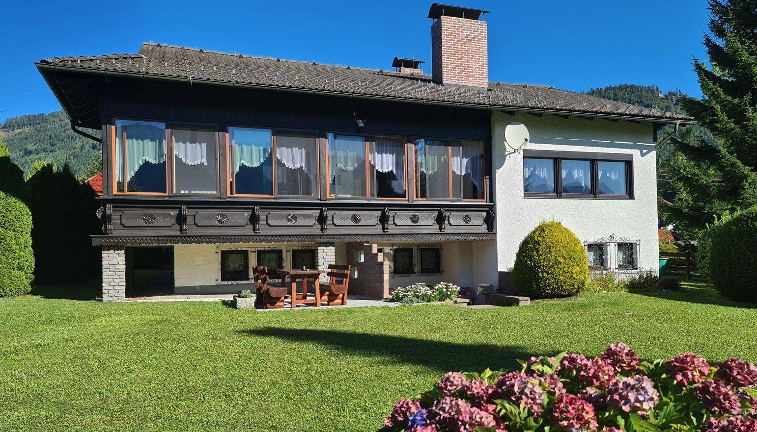 Traditionelles Haus mit Holzbalkon und gepflegtem Garten im Vordergrund unter blauem Himmel.