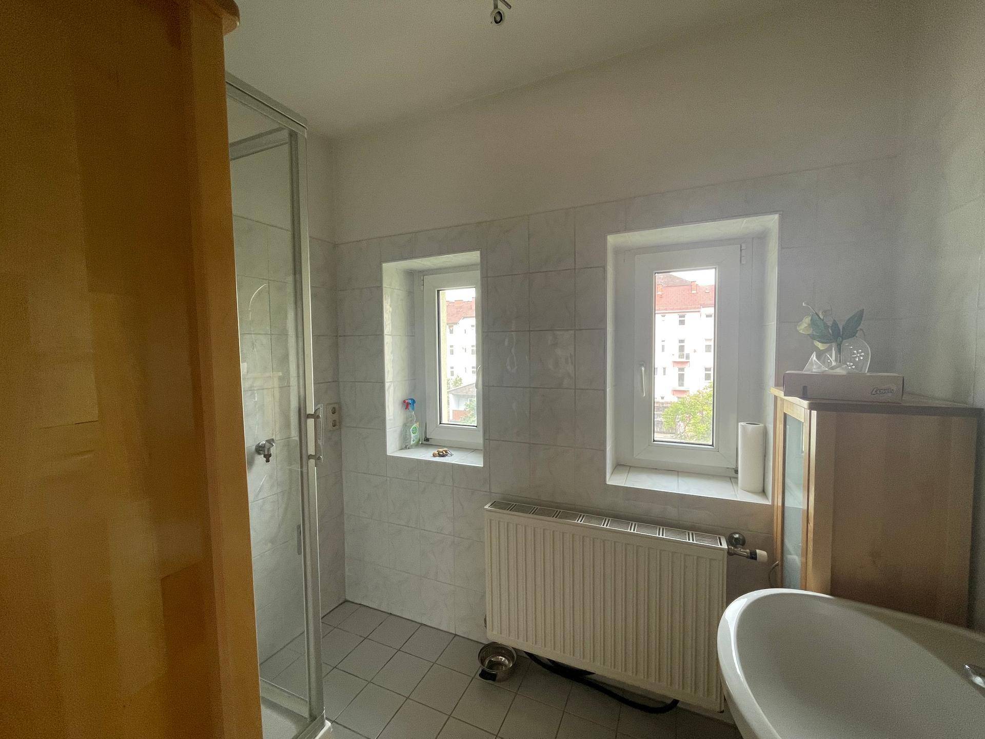 Badezimmer mit hellen Fliesen, Duschkabine und zwei Fenstern für Tageslicht.