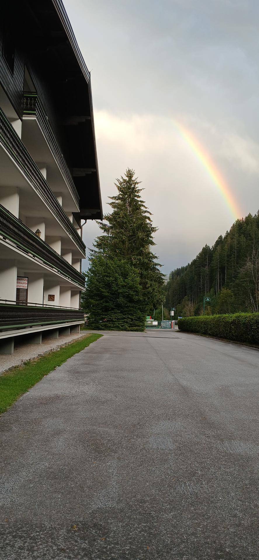 Gebäude mit Balkonen und Blick auf bewaldete Berge, unter einem Himmel mit Regenbogen.