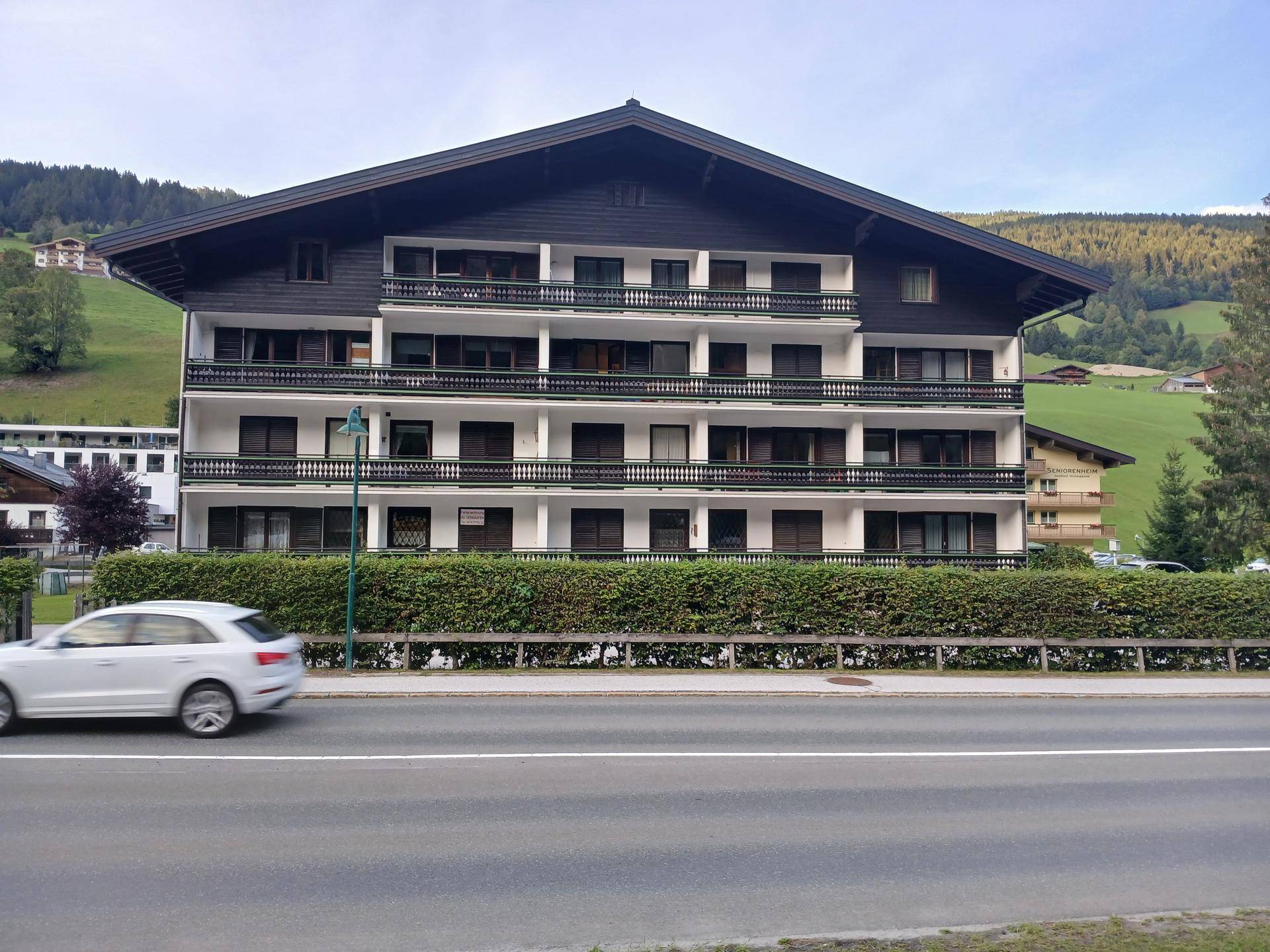 Mehrfamilienhaus im alpinen Stil mit dunklem Dach, vielen Balkonen und Blick auf grüne Berghänge.