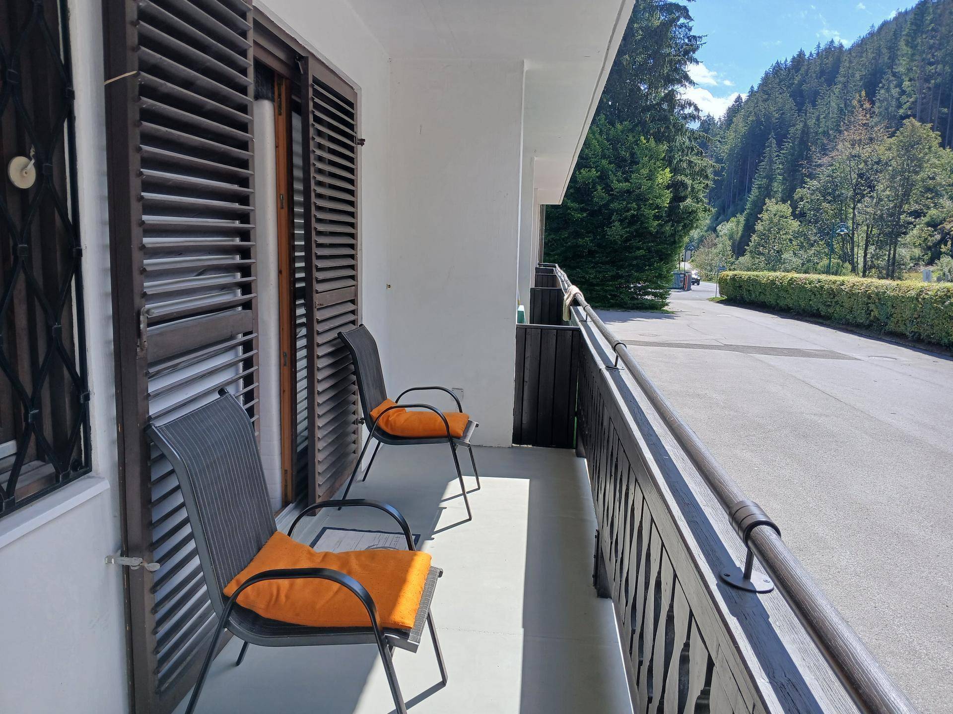 Sonniger Balkon mit zwei Stühlen und Blick auf bewaldete Berge und die Straße.