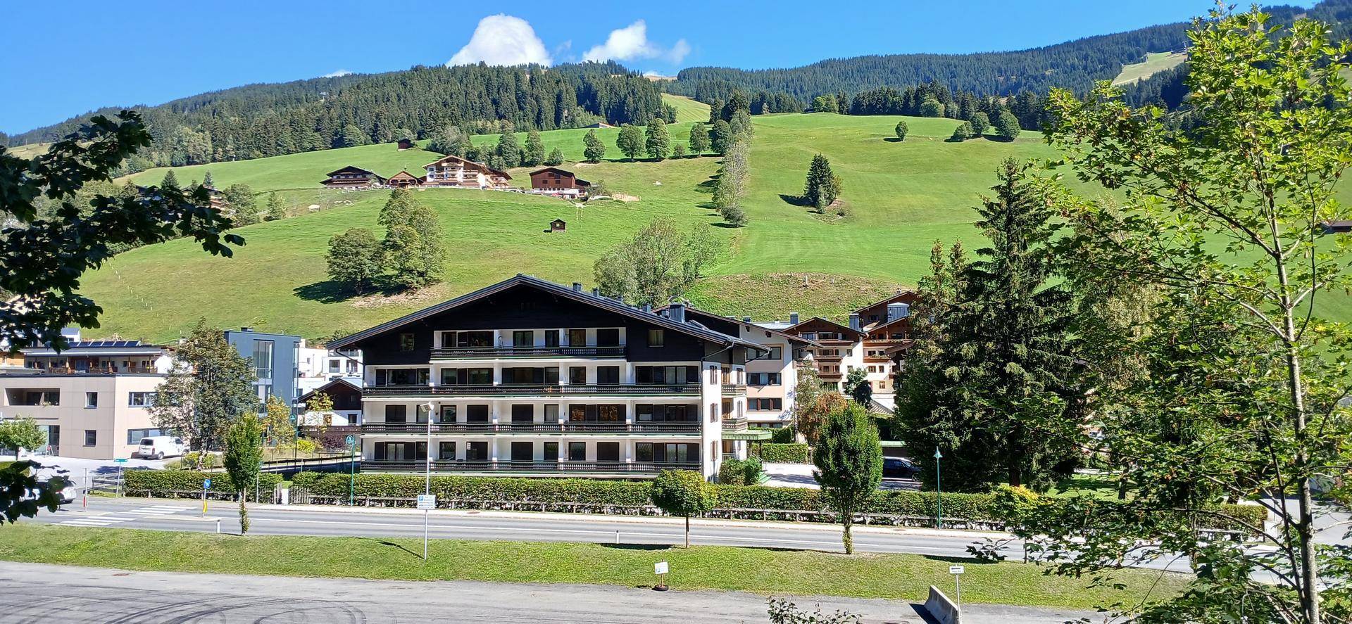 Mehrstöckiges Gebäude mit Balkonen vor einer malerischen grünen Berglandschaft mit weiteren Häusern.