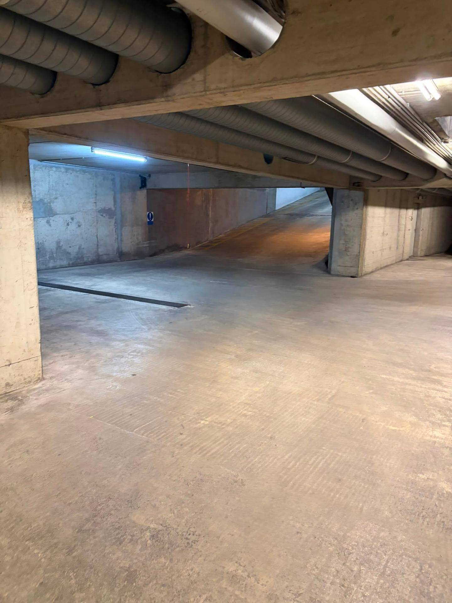 Innenansicht einer Tiefgarage mit sichtbaren Rohren an der Decke und einer Rampe im Hintergrund.