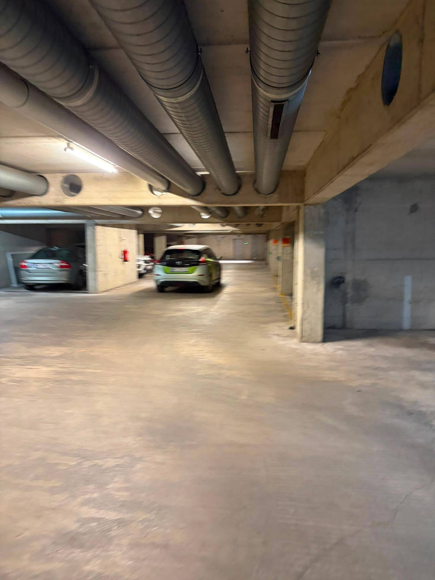 Geräumige Tiefgarage mit mehreren geparkten Fahrzeugen und sichtbaren Versorgungsleitungen an der Decke.
