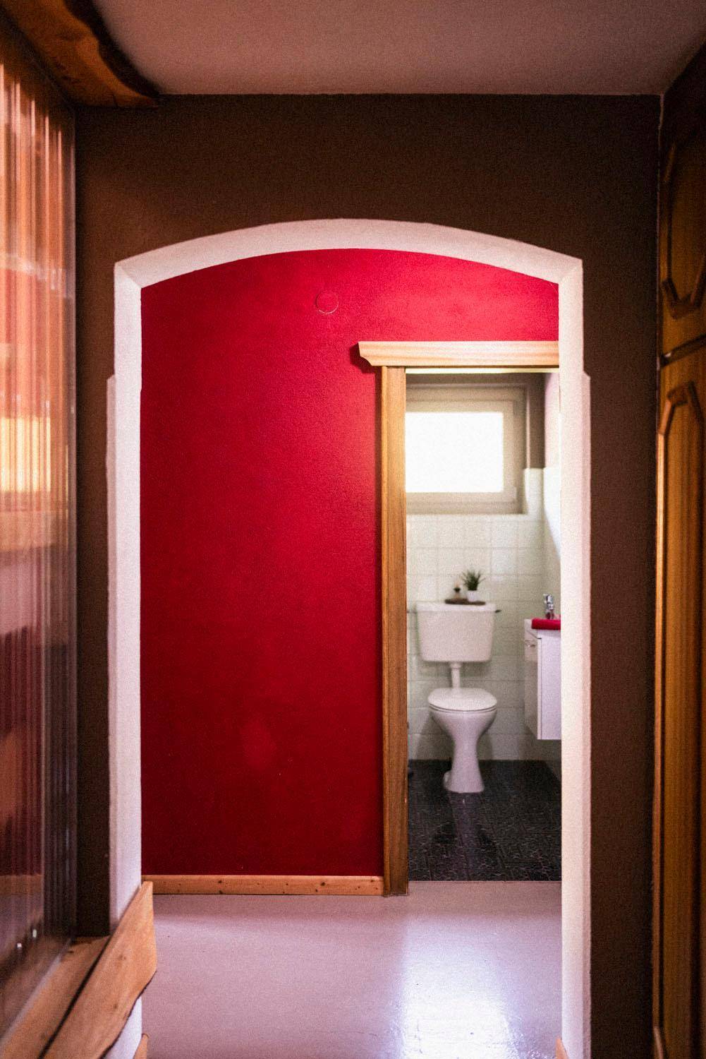 Flur mit roter Wand und Holztür, die zu einem kleinen WC mit Fenster führt.
