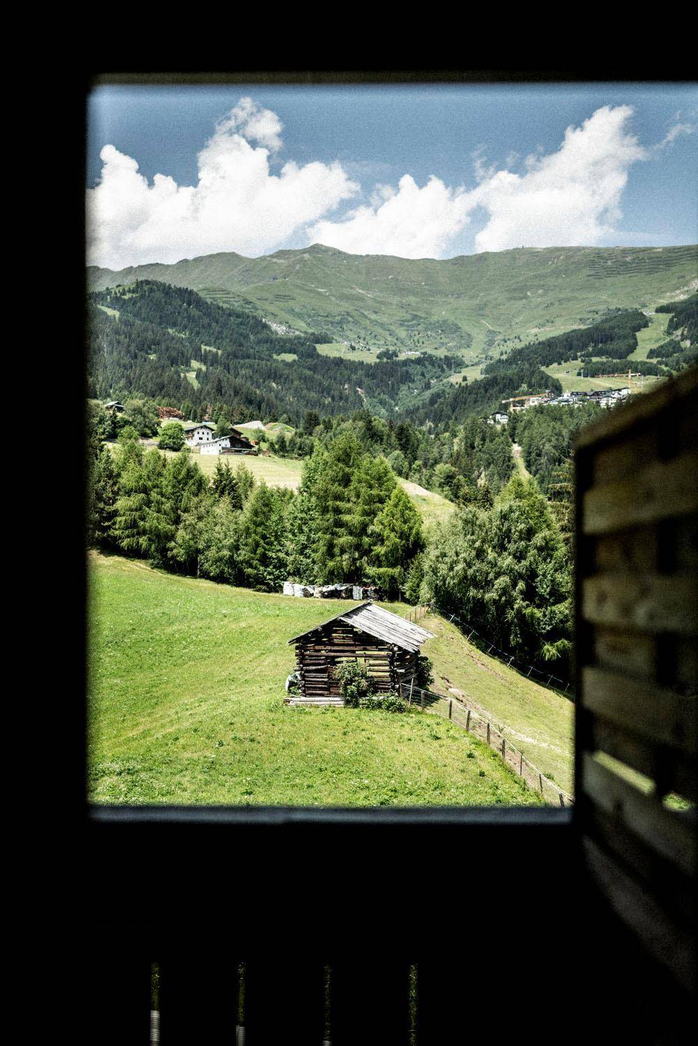 Malerischer Blick aus einem Fenster auf eine grüne Berglandschaft mit einem kleinen Holzhaus.