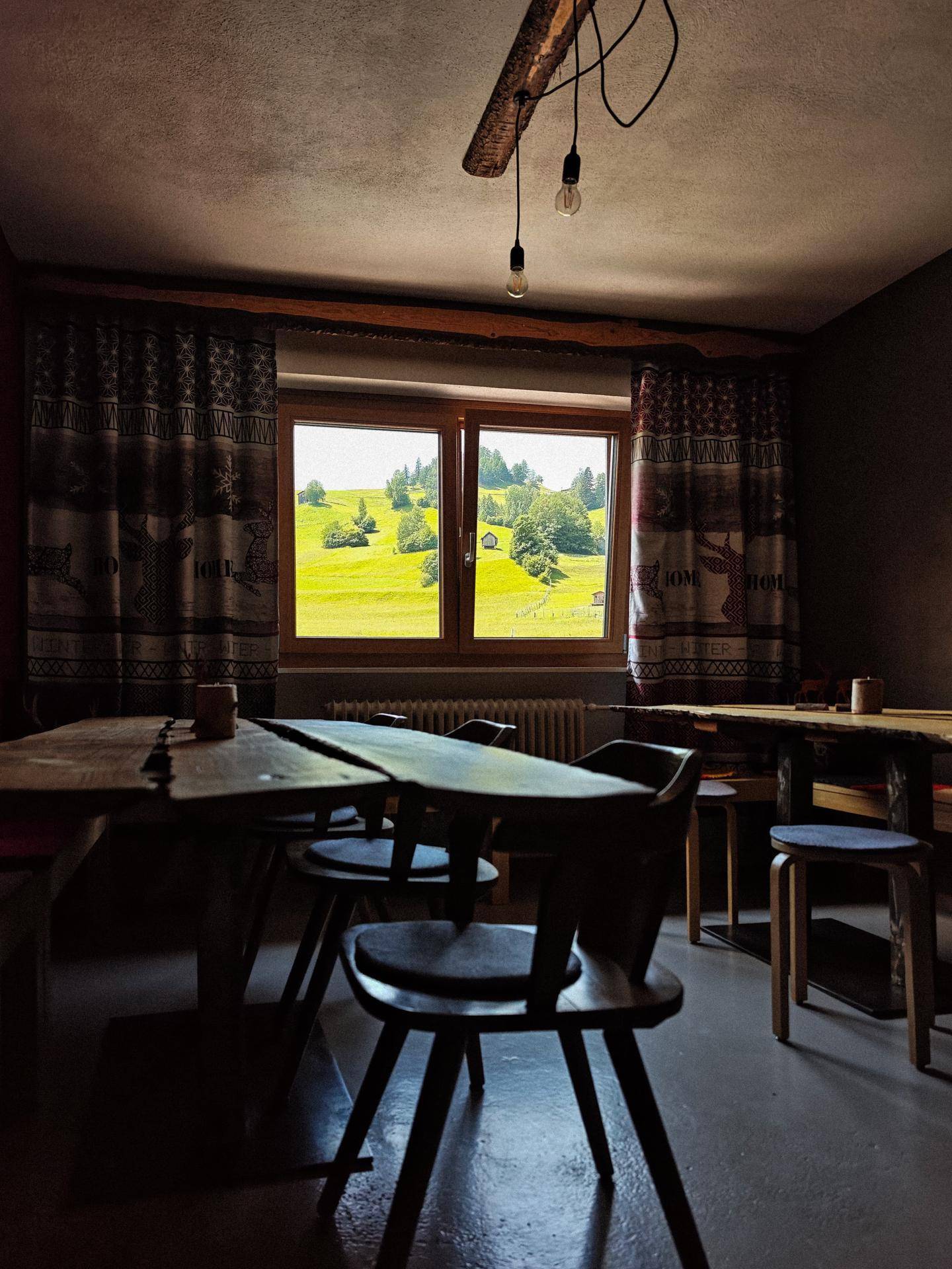 Rustikaler Essbereich mit Holztisch und Stühlen, einem Fenster mit Blick auf eine grüne Wiese und sichtbaren Deckenbalken.