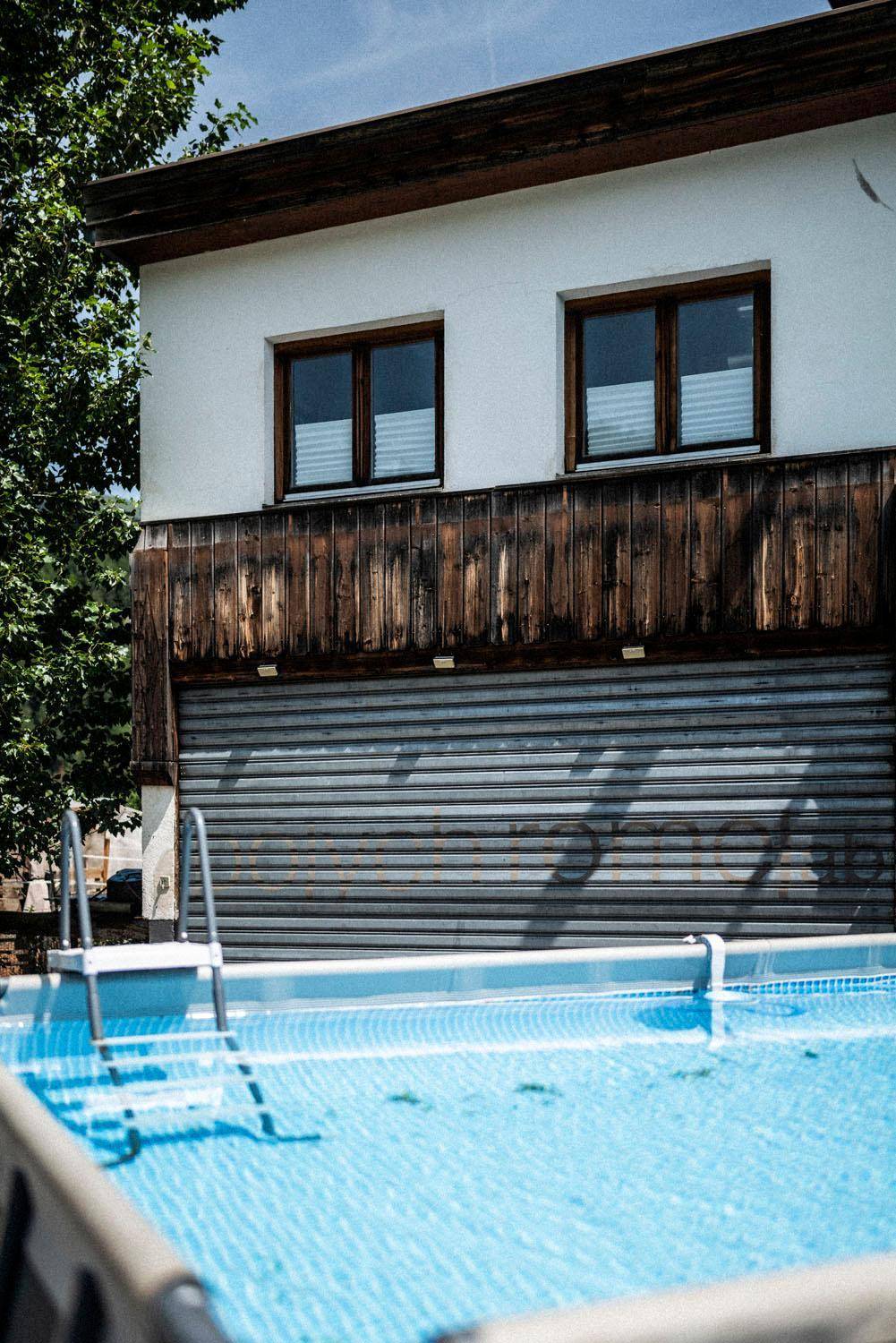 Außenbereich mit oberirdischem Pool vor einem Gebäude mit Holzfassade und Rollladen.