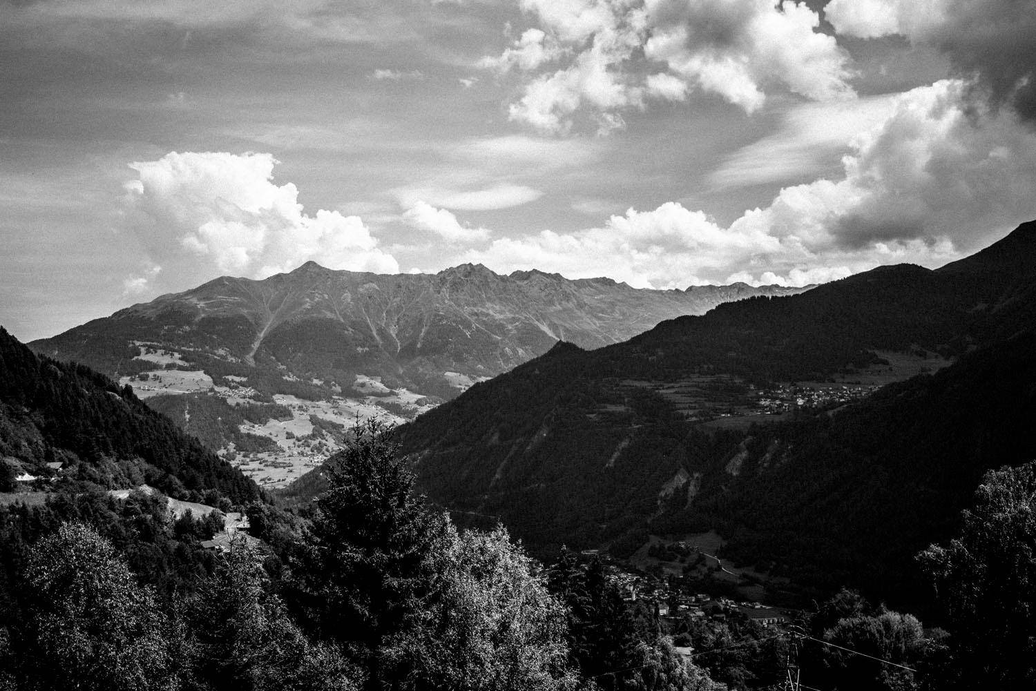 Weitläufige Schwarz-Weiß-Ansicht einer bergigen Landschaft mit bewaldeten Tälern und Wolken am Himmel.