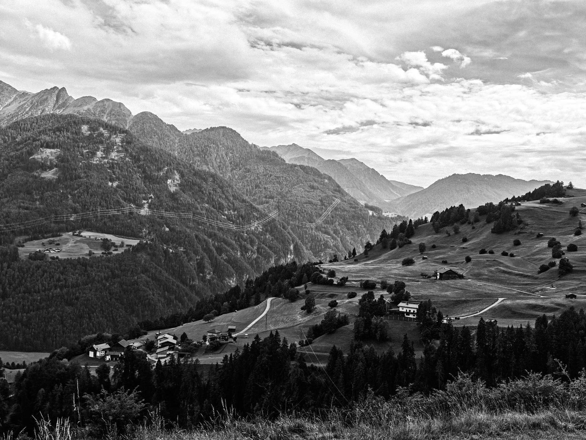 Schwarz-Weiß-Aufnahme einer weiten Berglandschaft mit bewaldeten Hängen und vereinzelten Häusern.