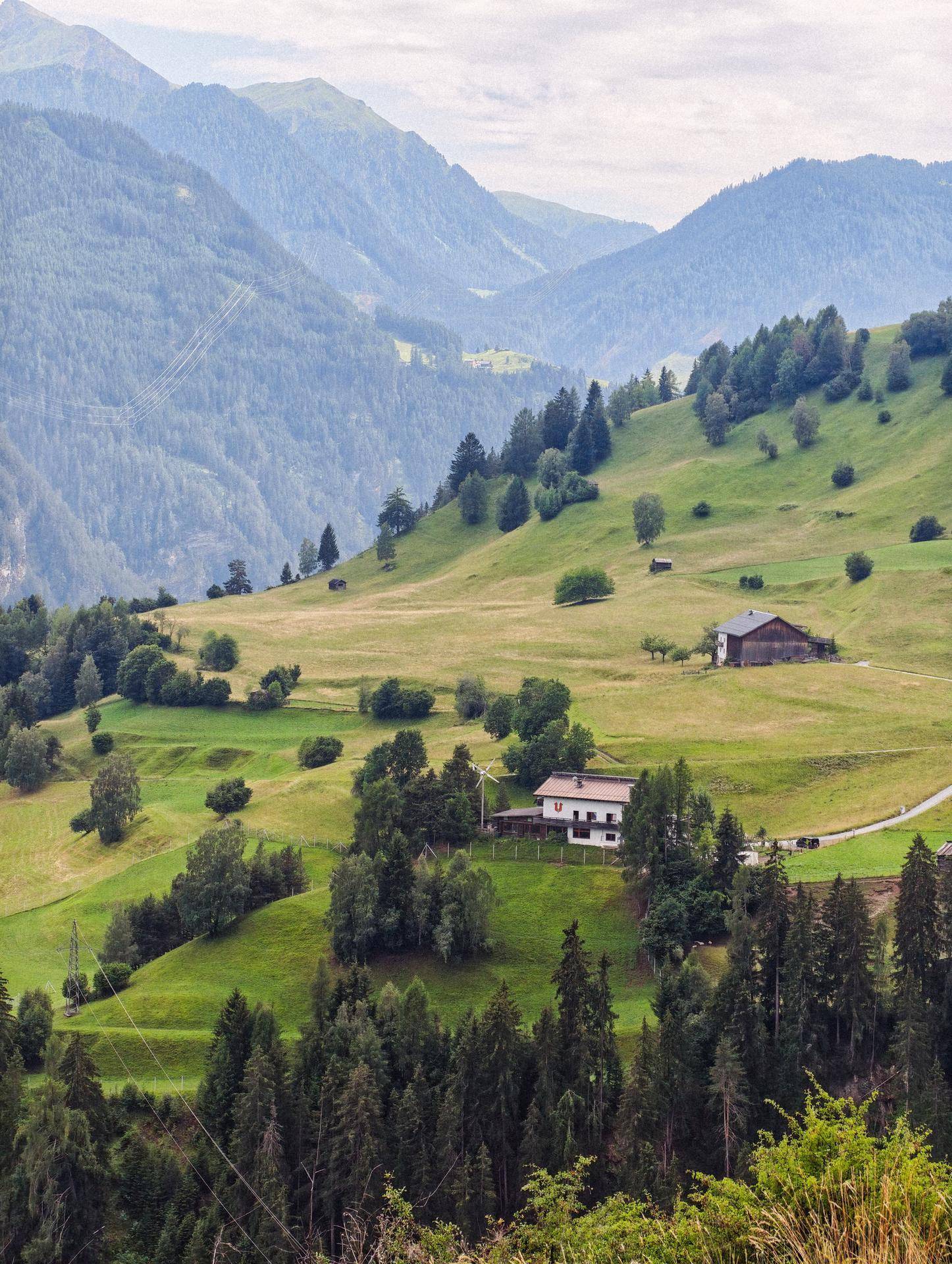 Grüne Berglandschaft mit sanften Hügeln, Wäldern und einem einzelnen Haus in der Ferne unter bewölktem Himmel.