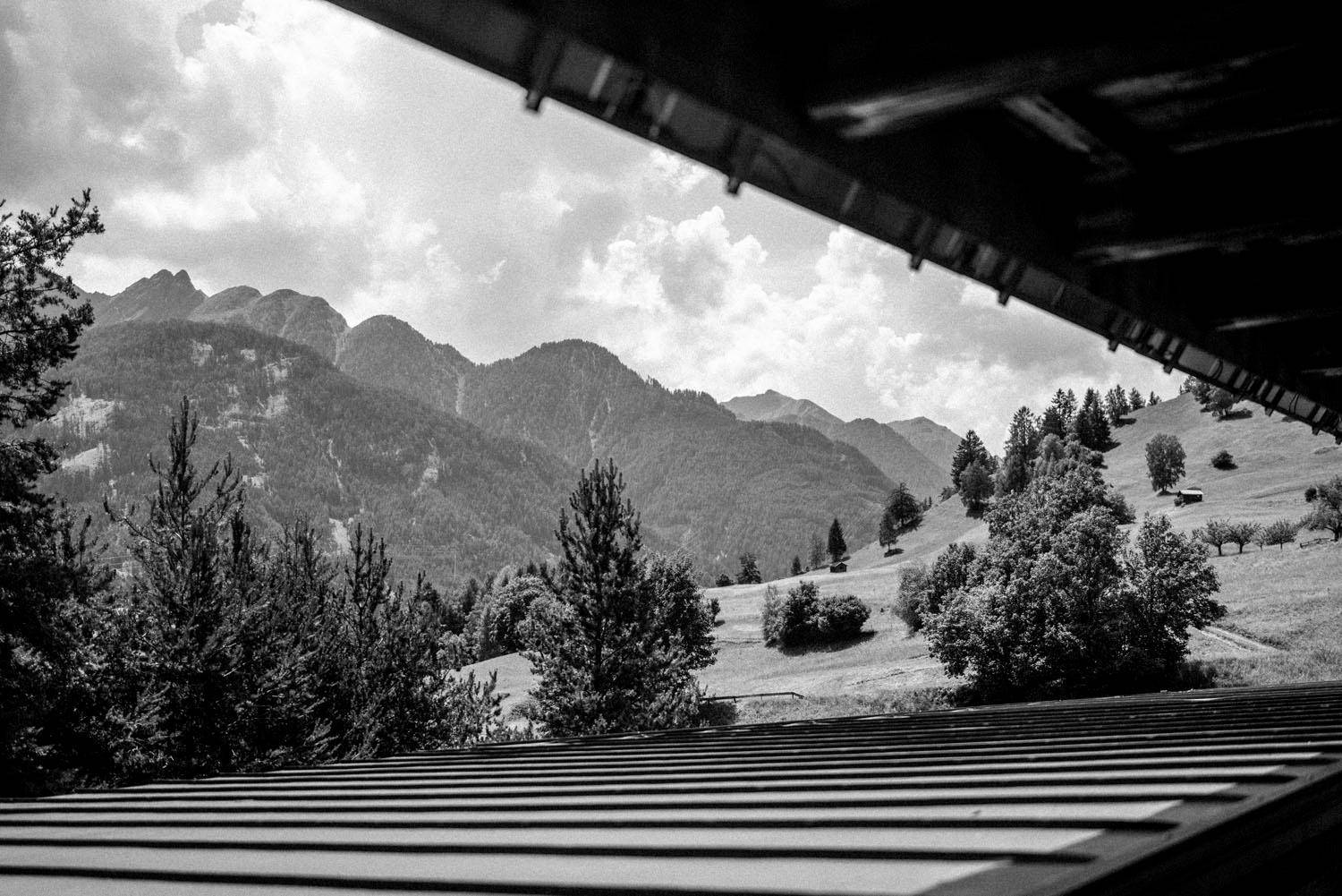 Schwarz-Weiß-Blick von einem überdachten Bereich auf eine weite Berglandschaft mit Bäumen und Wiesen.