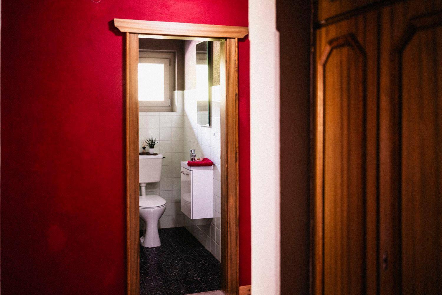 Flur mit roter Wand und Holztür, die zu einem kleinen WC mit Fenster und modernem Waschtisch führt.