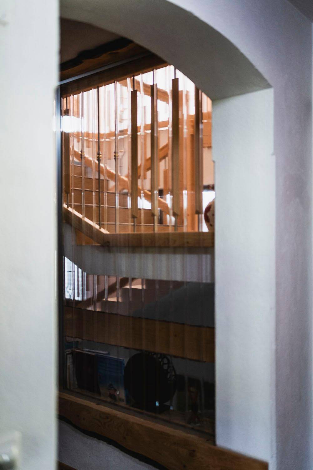 Blick durch einen Torbogen auf ein helles Treppenhaus mit Holzgeländer und viel Tageslicht.