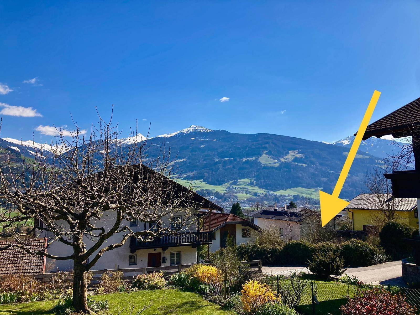 Weitläufiger Ausblick auf eine Wohnsiedlung mit grünen Gärten und beeindruckenden Bergen unter strahlend blauem Himmel.
