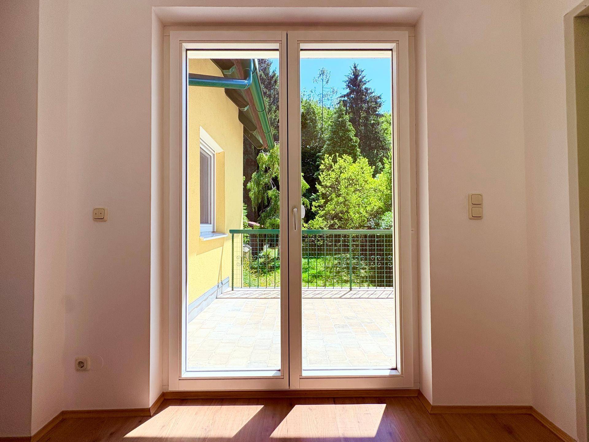 Innenraum mit großen Fenstertüren, die den Blick auf einen Balkon und die Natur freigeben.