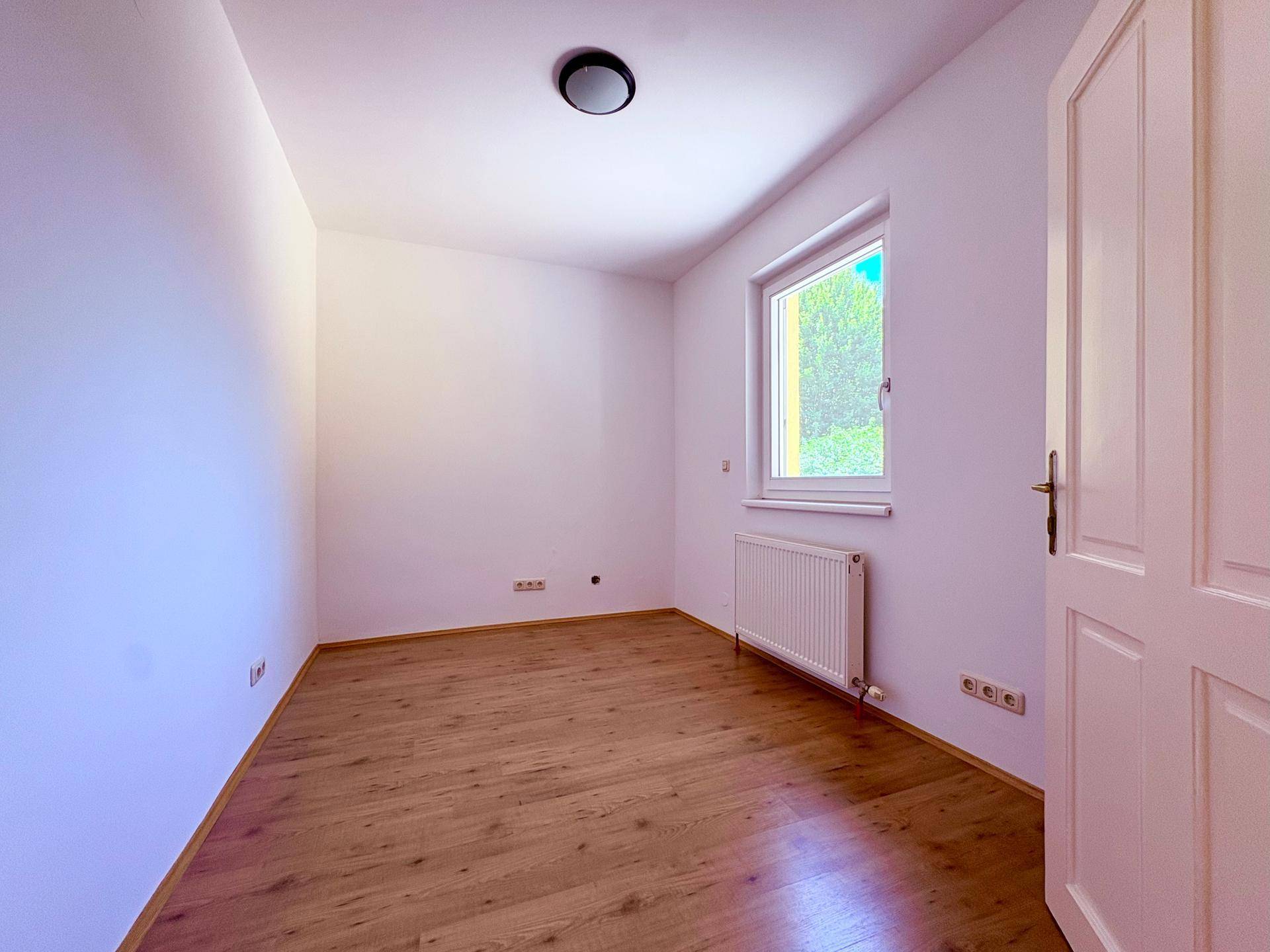 Helles Schlafzimmer mit Holzboden, weiß gestrichenen Wänden und einem Fenster mit Blick ins Grüne.