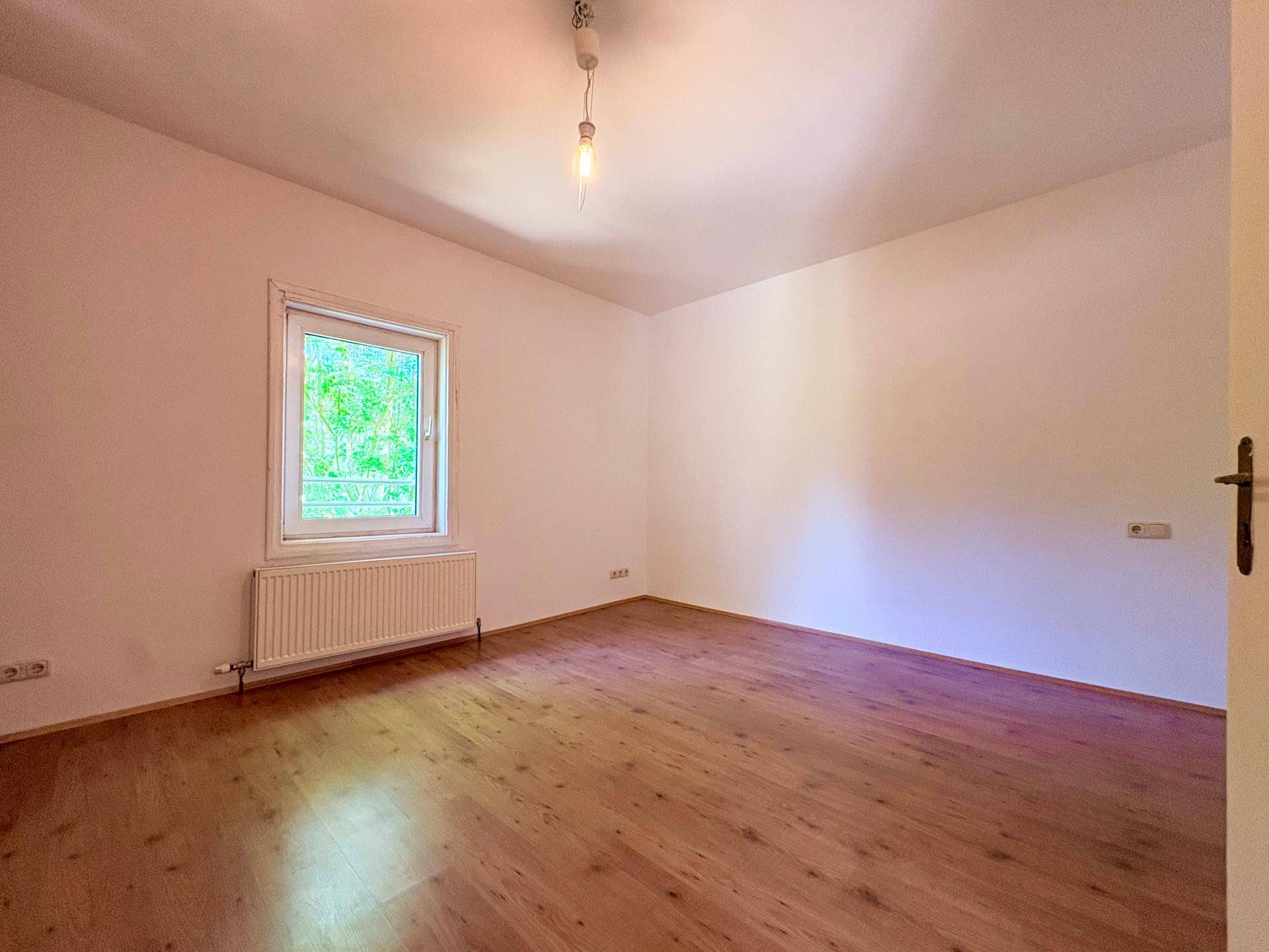Geräumiges Schlafzimmer mit Holzboden, weißen Wänden und einem Fenster mit Blick ins Grüne.