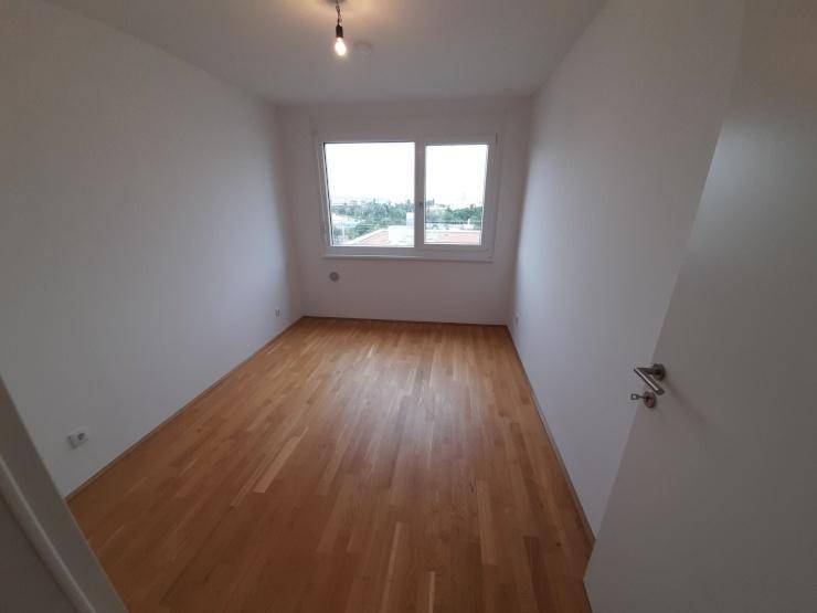 Leeres Zimmer mit hellem Holzboden und einem Fenster, das viel Tageslicht hereinlässt.