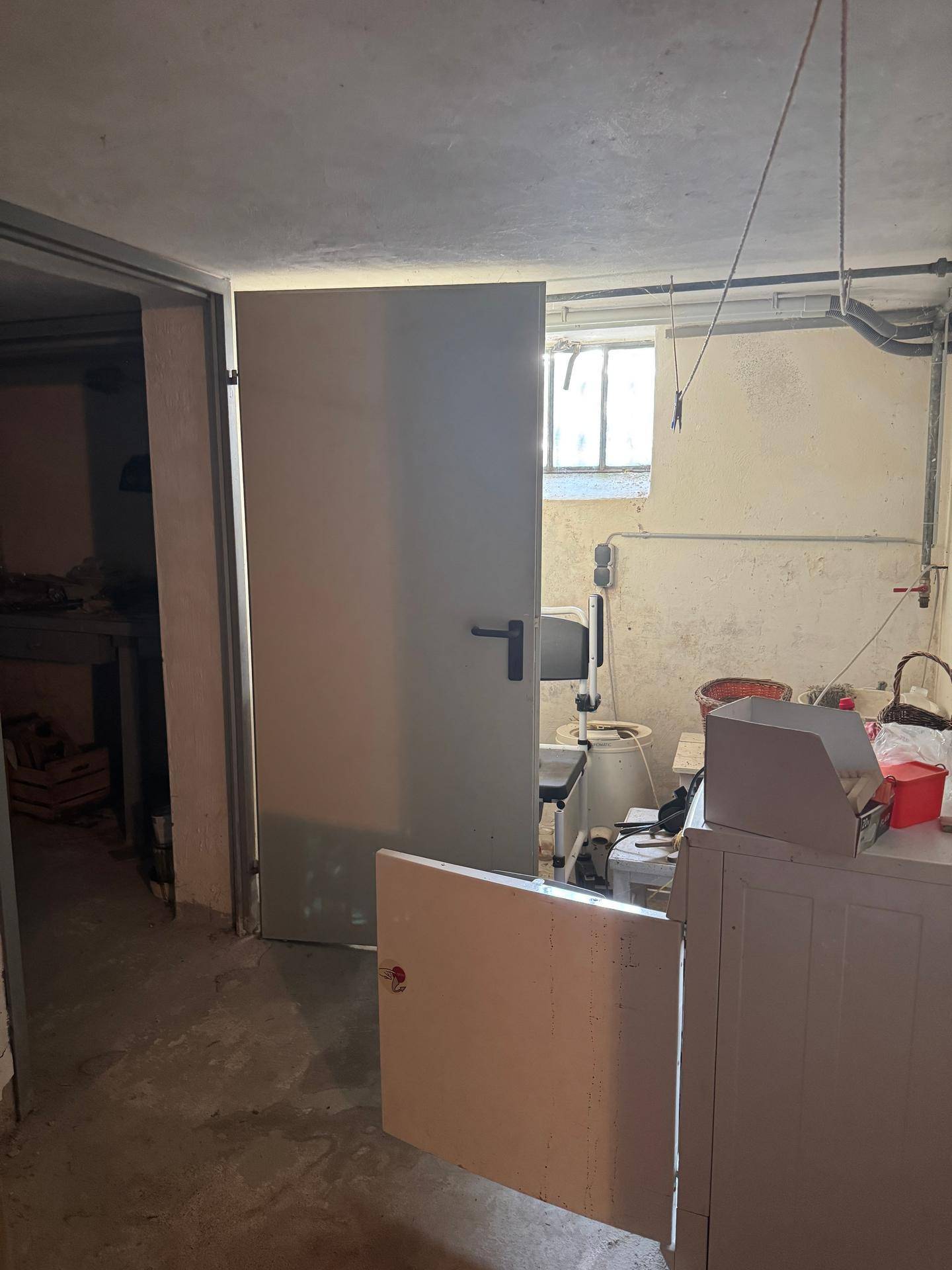Kellerraum mit einer Tür, kleinem Fenster und sichtbaren Rohren an der Decke, unfertiger Zustand.
