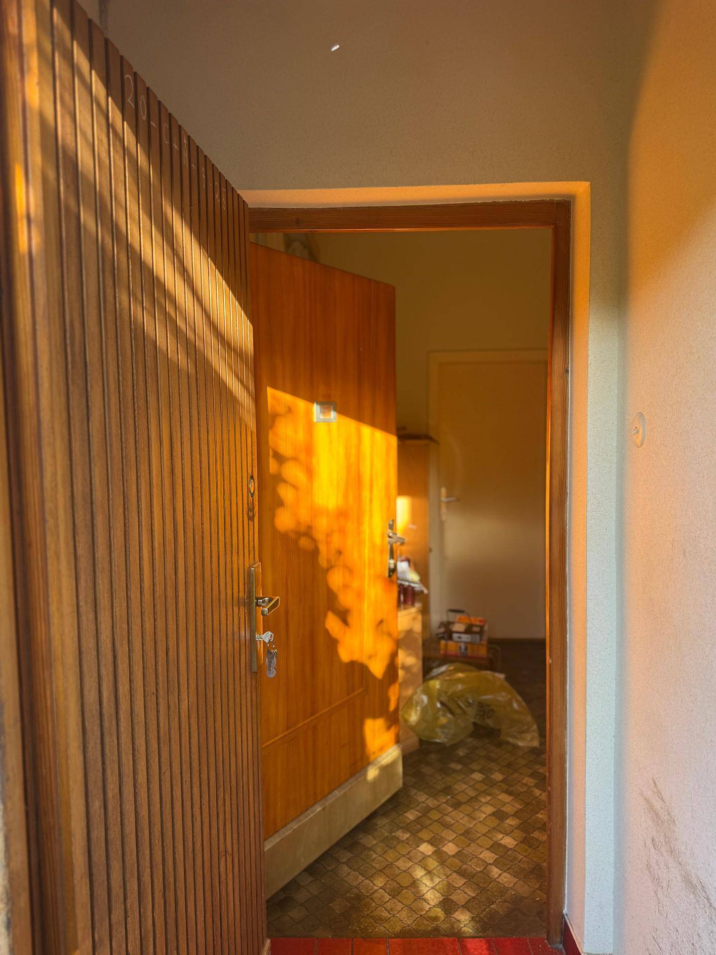 Eingangsbereich mit offener Holztür und Fliesenboden, beleuchtet von warmem Sonnenlicht.