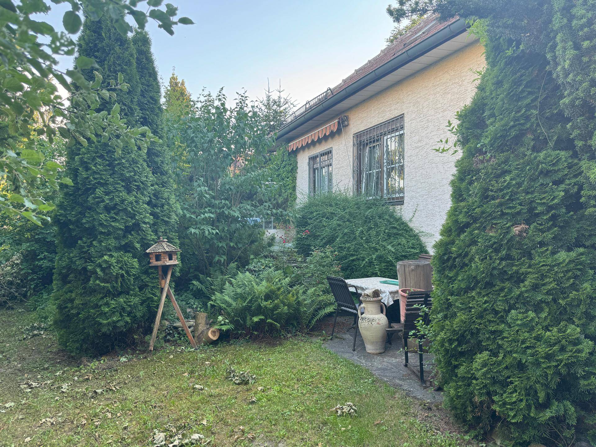 Garten mit Terrasse und Sitzgelegenheiten neben dem Haus, umgeben von hohen Hecken und Bäumen.