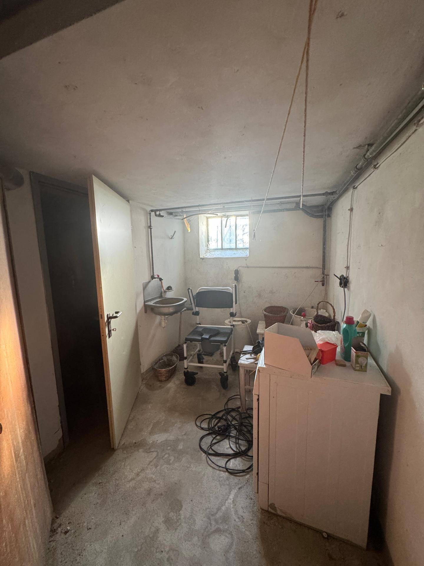 Kellerraum mit Waschbecken, kleinem Fenster und verschiedenen Gegenständen, in ungenutztem Zustand.