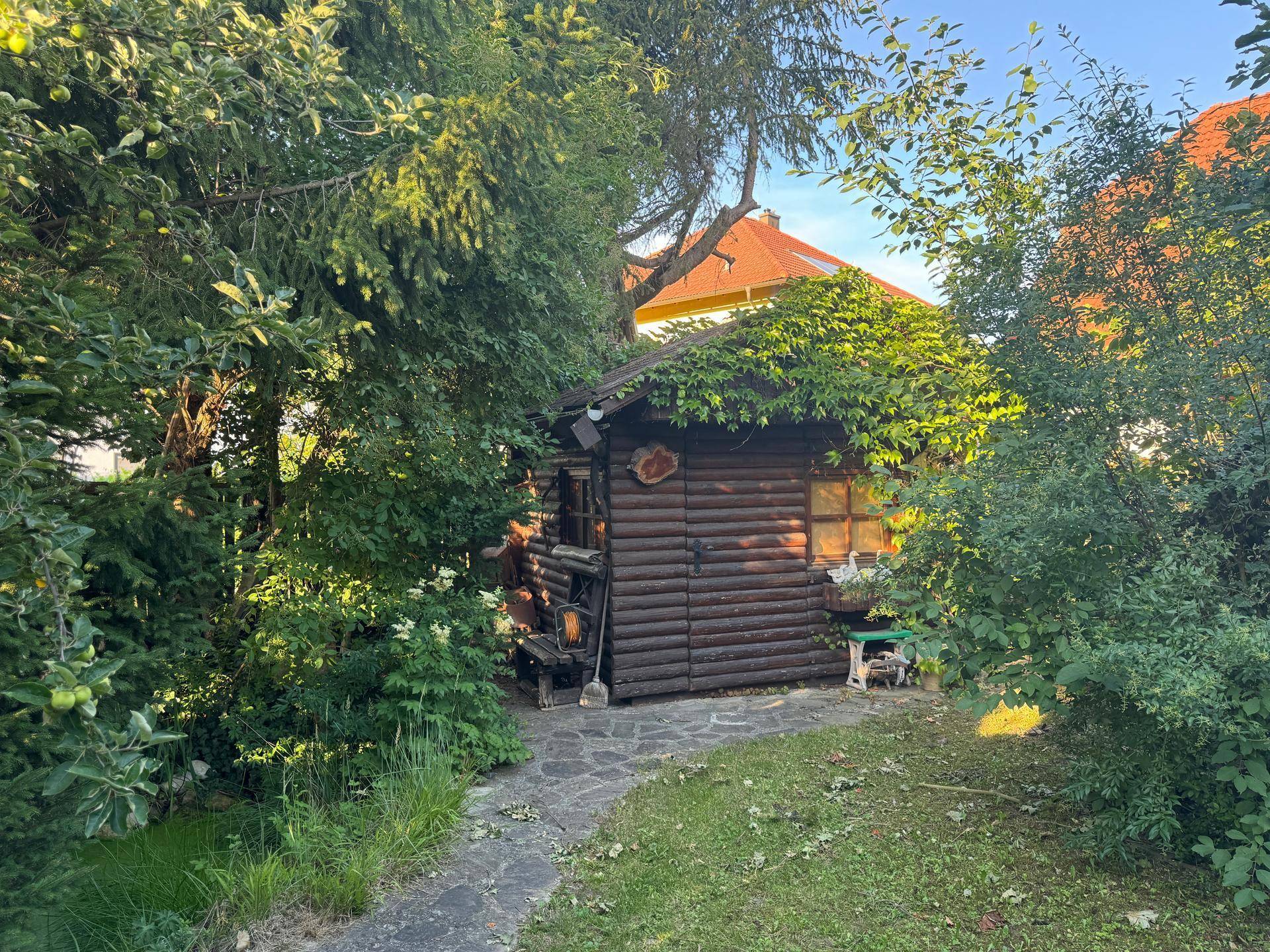 Rustikales Holzgartenhaus umgeben von Bäumen und Sträuchern, mit einem kleinen Steinweg davor.