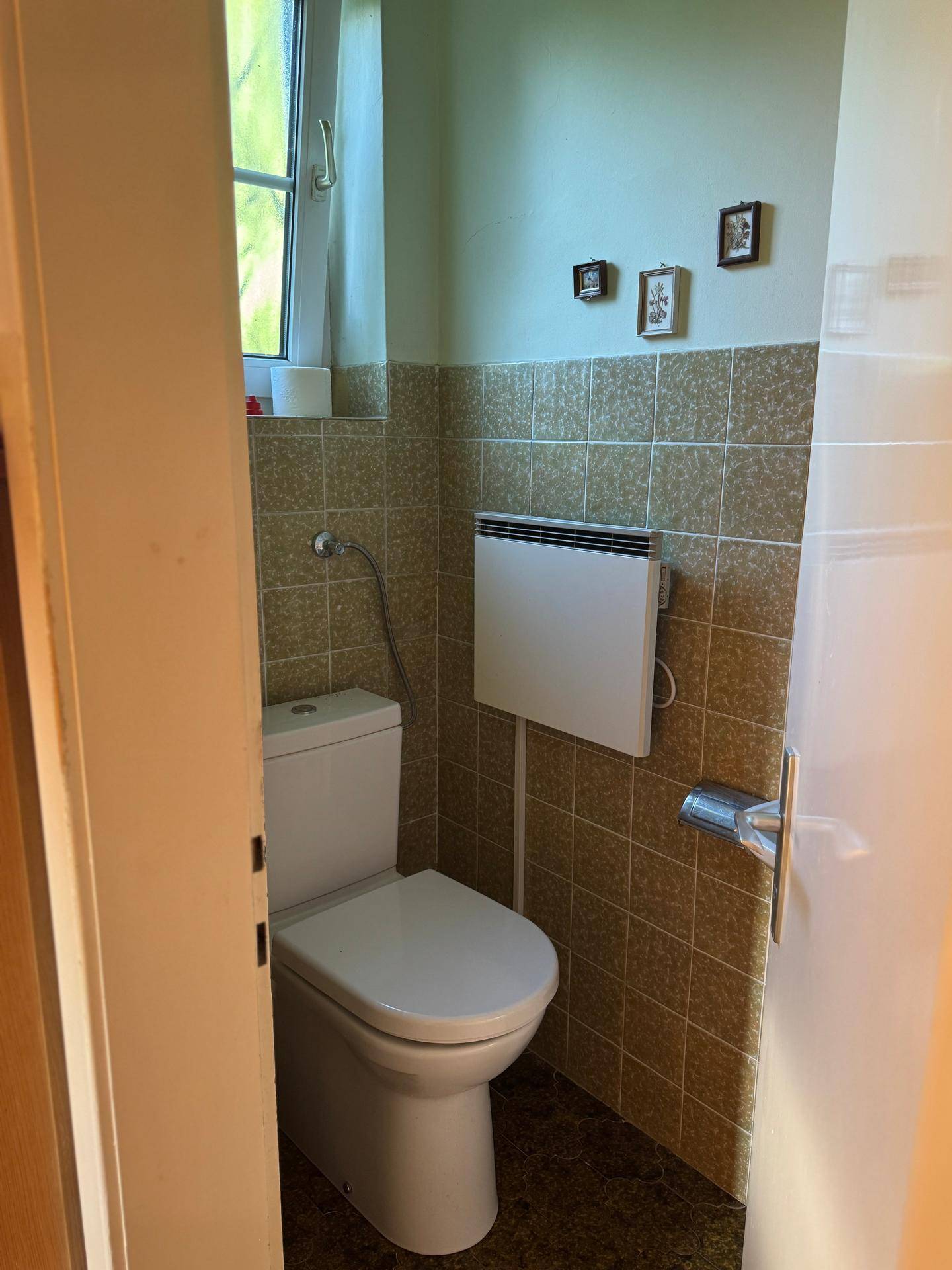 Kleines WC mit beigefarbenen Fliesen, Fenster und einem Wandheizkörper, in einfachem Zustand.