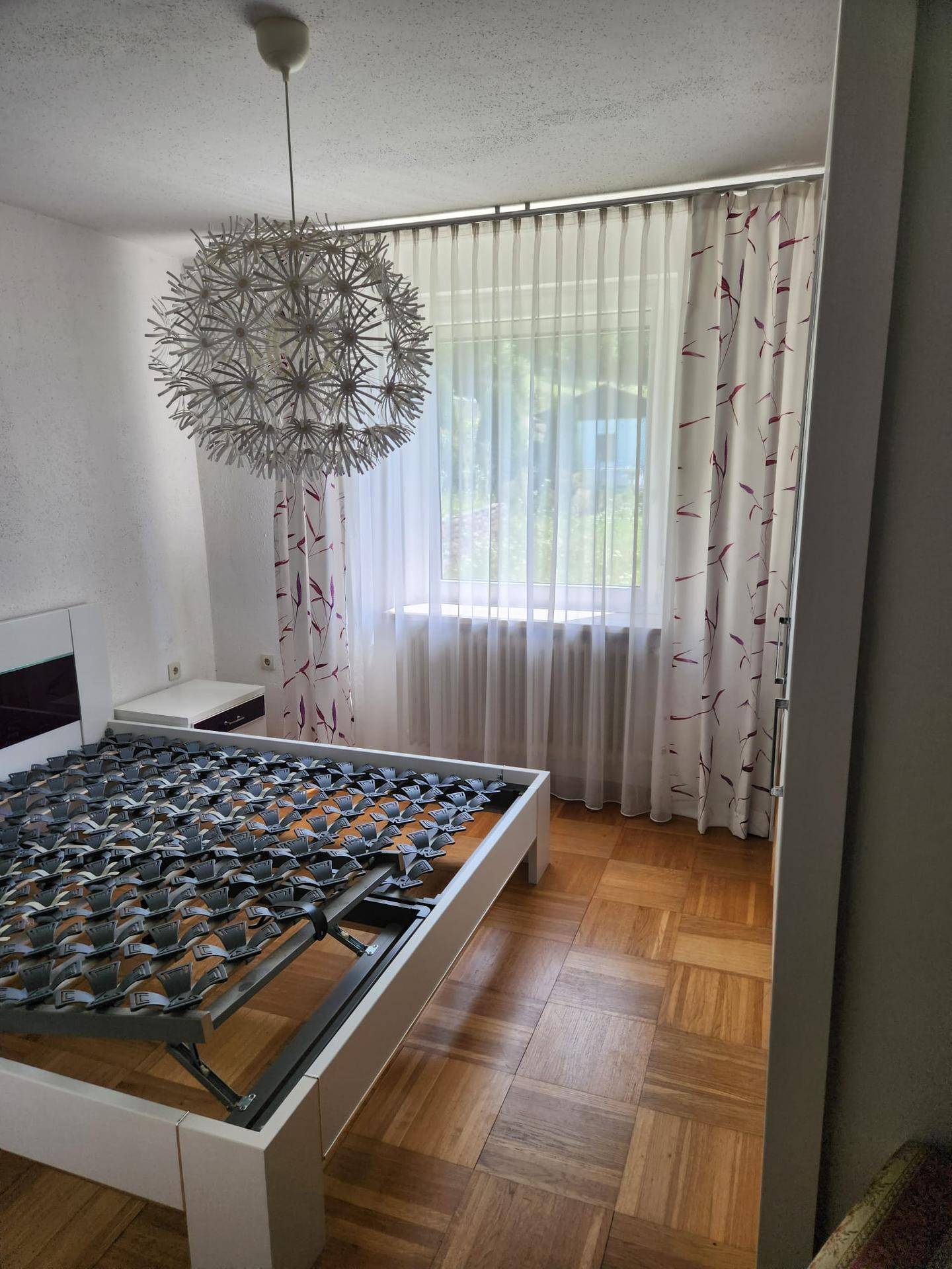 Schlafzimmer mit Parkettboden, großem Fenster und dekorativer Deckenleuchte.