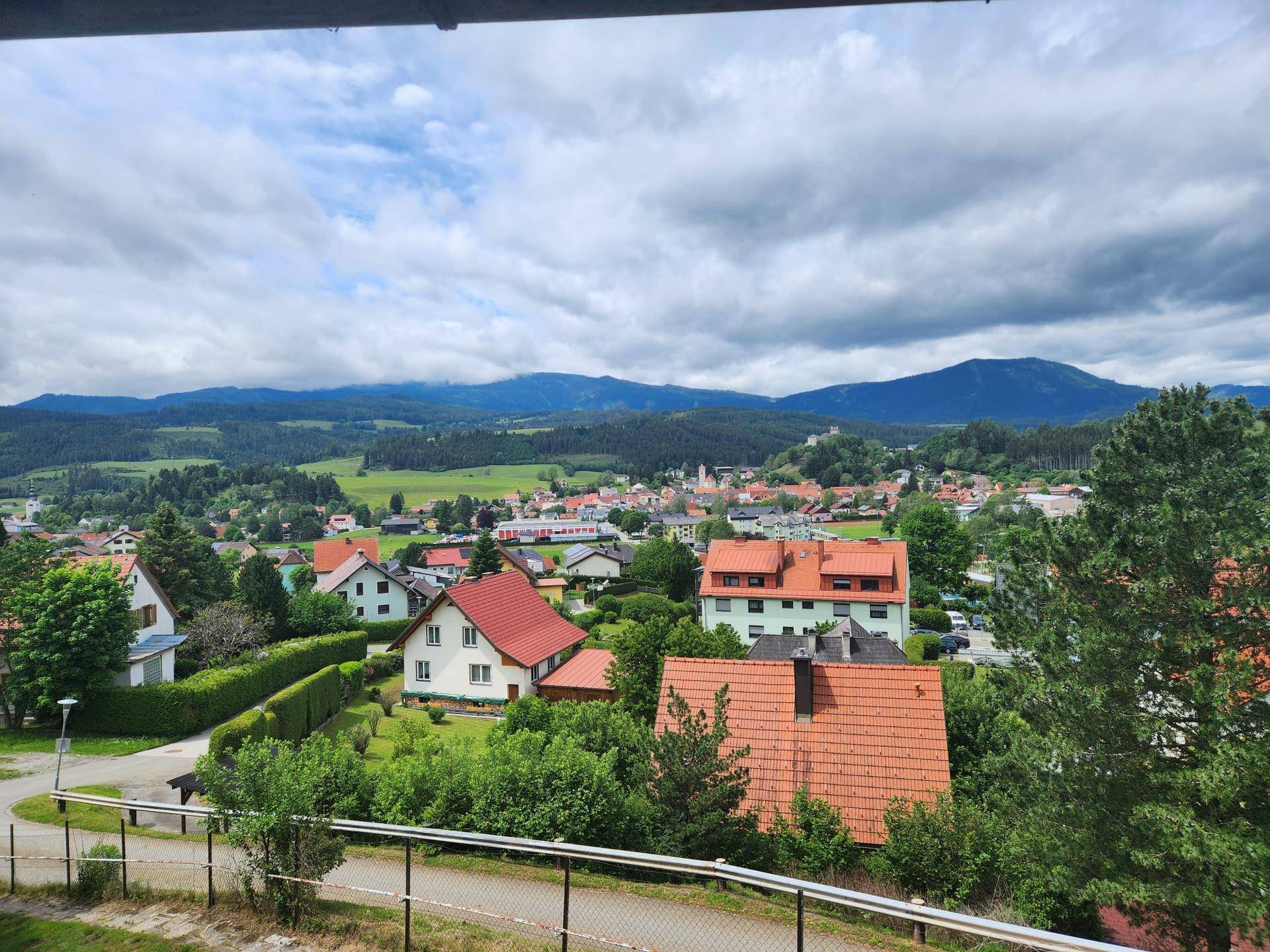 Weitläufiger Blick über eine malerische Stadt mit roten Dächern und einer Bergkulisse im Hintergrund.