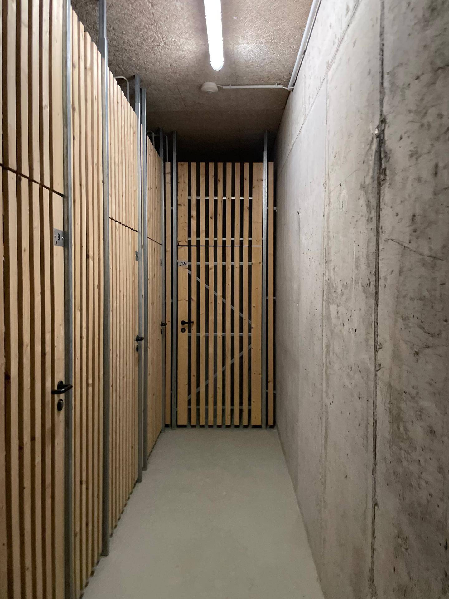 Moderner Kellergang mit Betonwänden und Holzlattenverschlägen für Lagerräume.