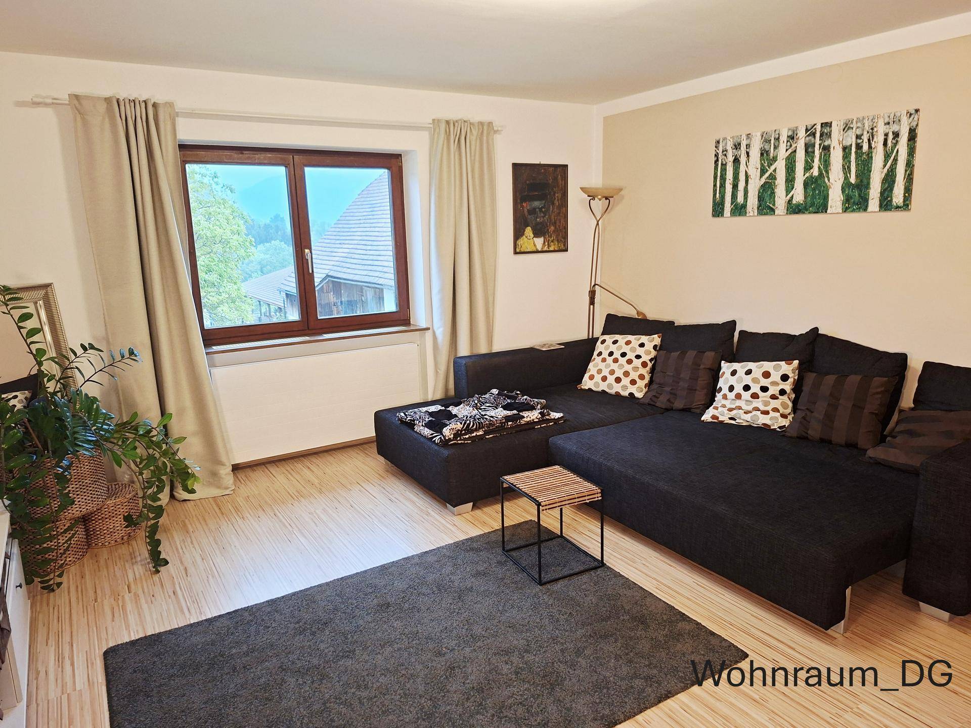 Gemütlicher Wohnbereich im Dachgeschoss mit dunklem Ecksofa, Teppich und Fenster mit Blick nach draußen.