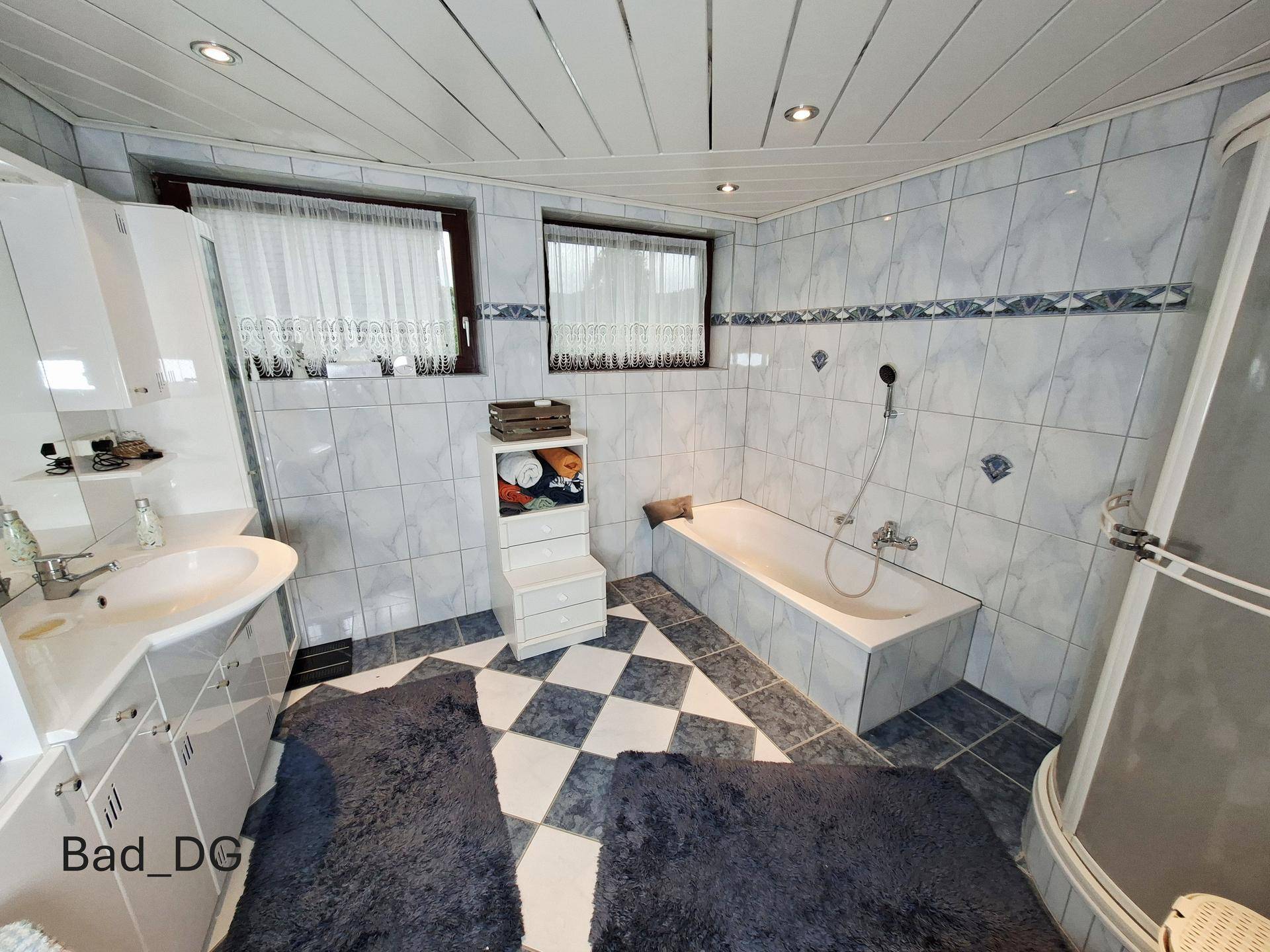 Badezimmer im Dachgeschoss mit blauen und weißen Fliesen, Badewanne, Duschkabine und zwei Fenstern.