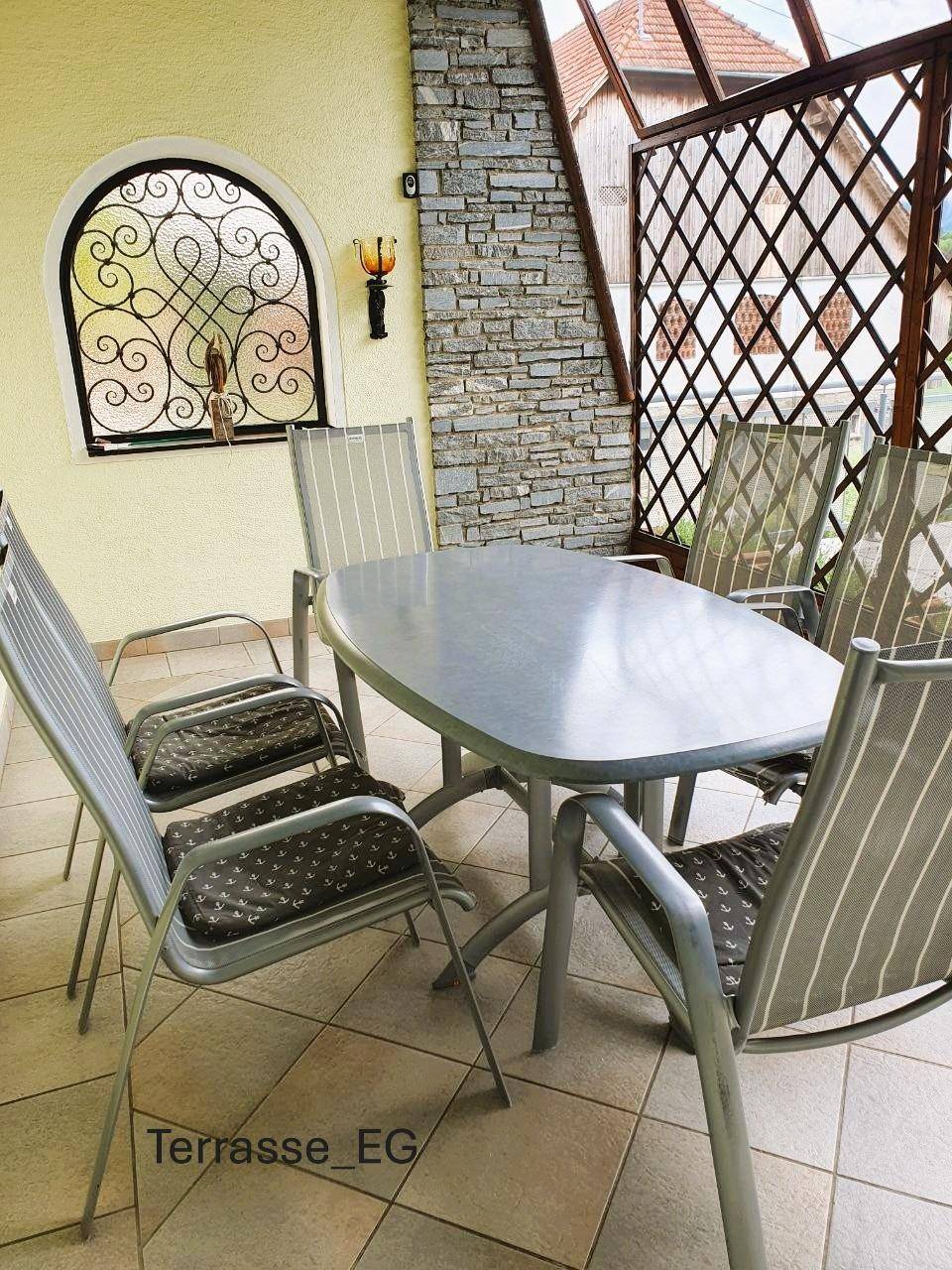 Überdachter Sitzbereich auf der Terrasse mit Tisch und Stühlen, umgeben von Stein- und Holzelementen.