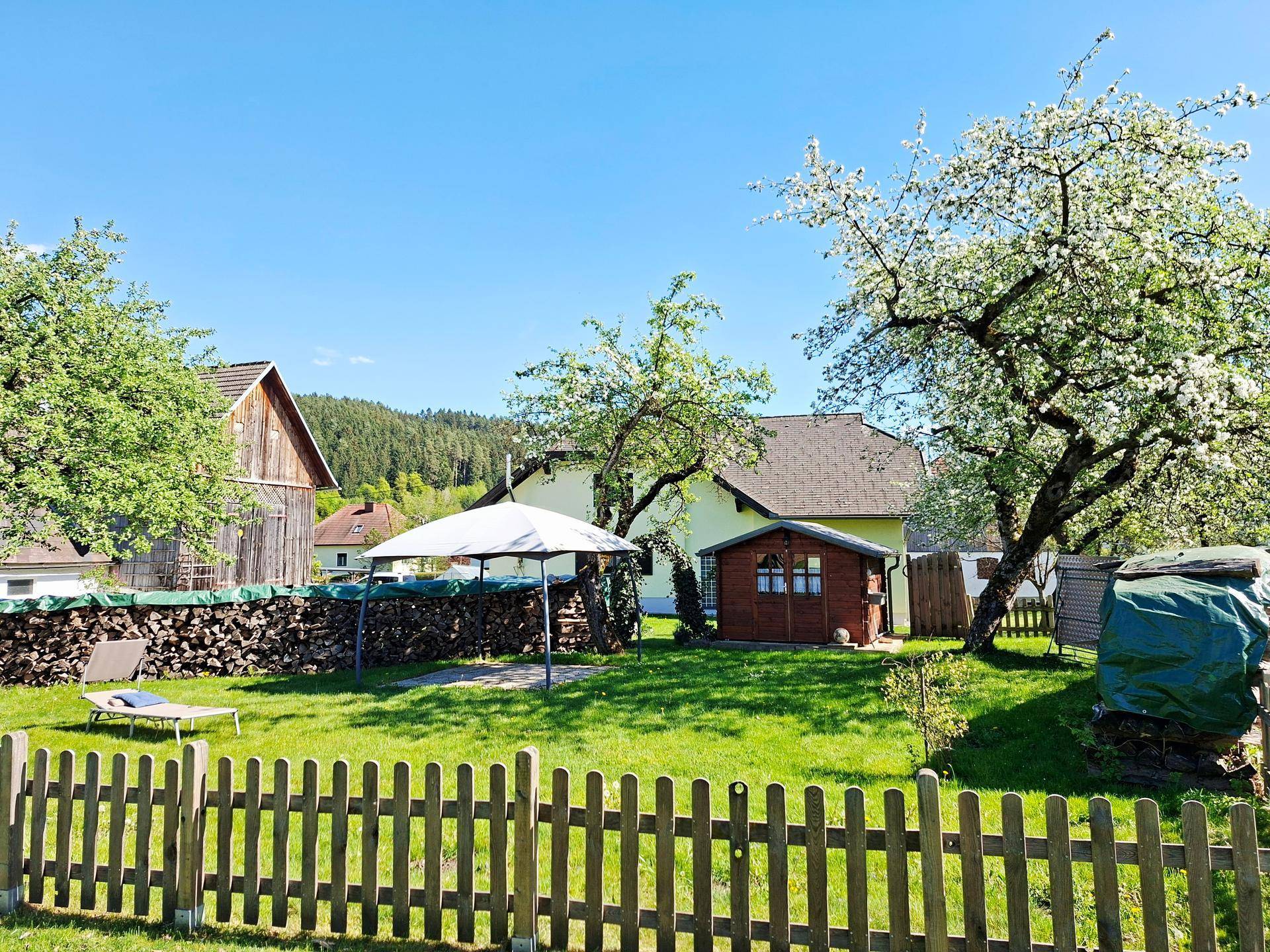Idyllischer Gartenbereich mit Gartenhaus, Holzstapel und schattenspendenden Bäumen, ideal zum Entspannen.