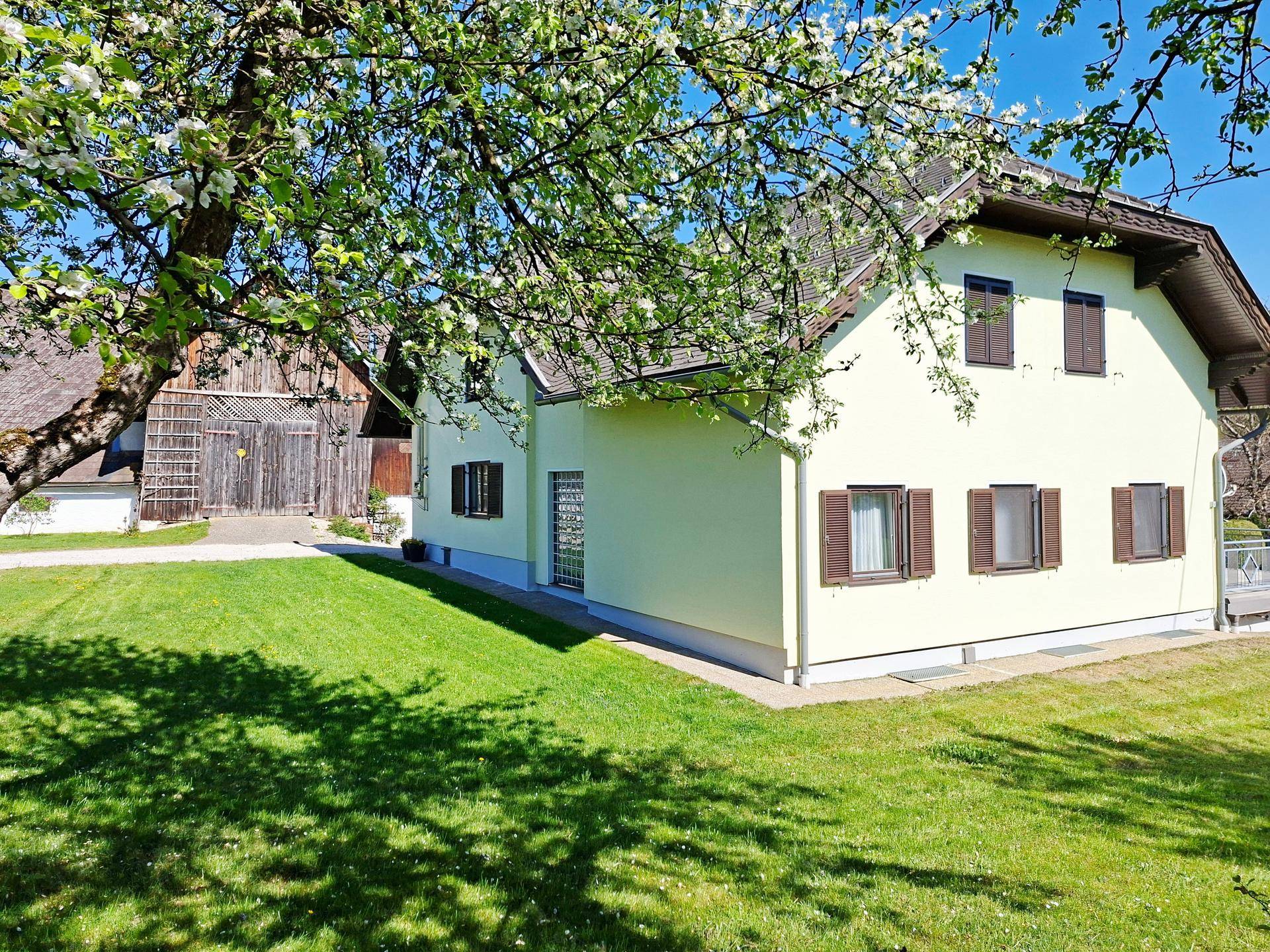Seitenansicht des Hauses mit gepflegtem Rasen und blühenden Bäumen unter strahlend blauem Himmel.