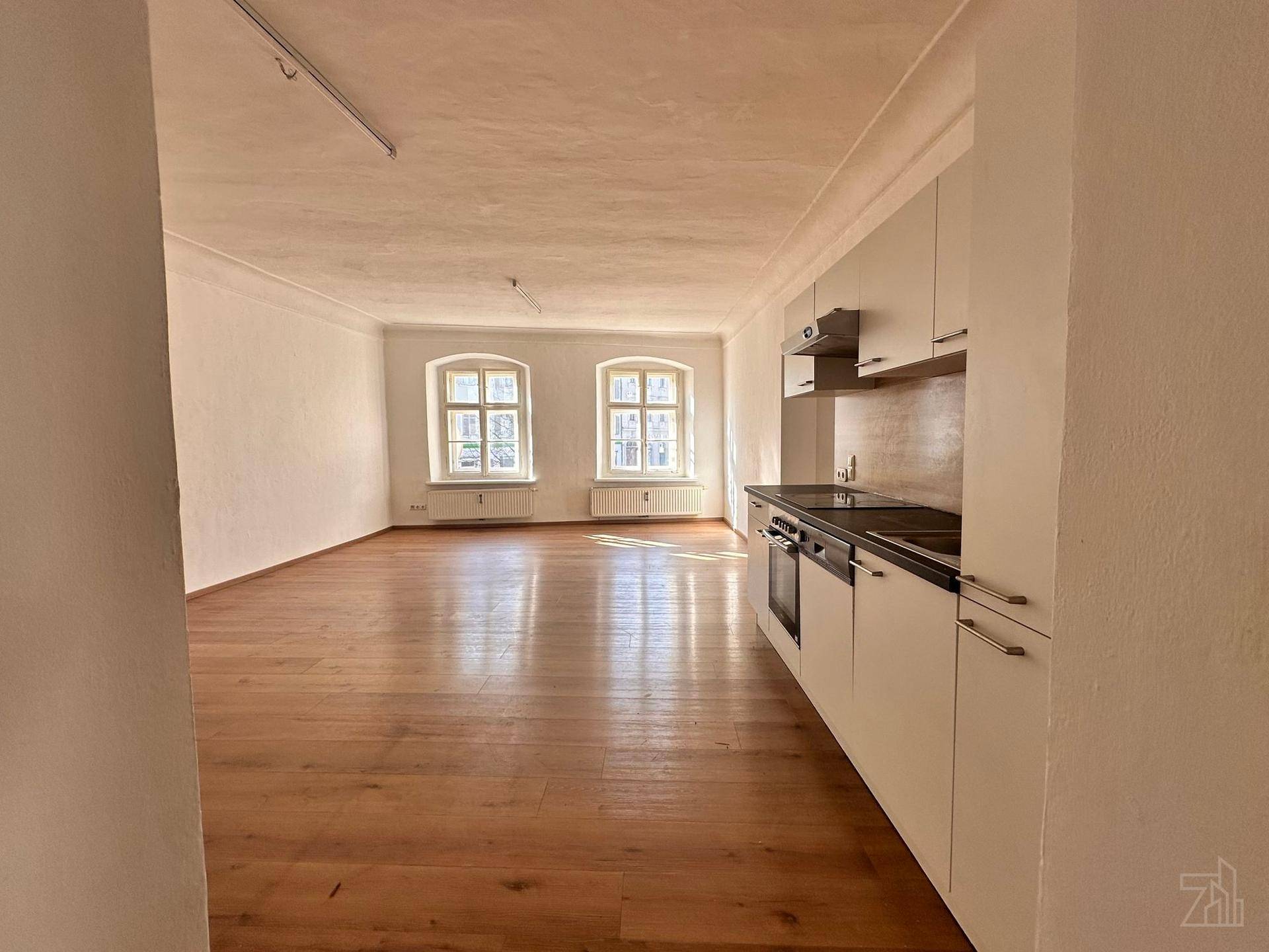 Heller Wohnbereich mit integrierter Küche, Holzboden und zwei großen Fenstern.