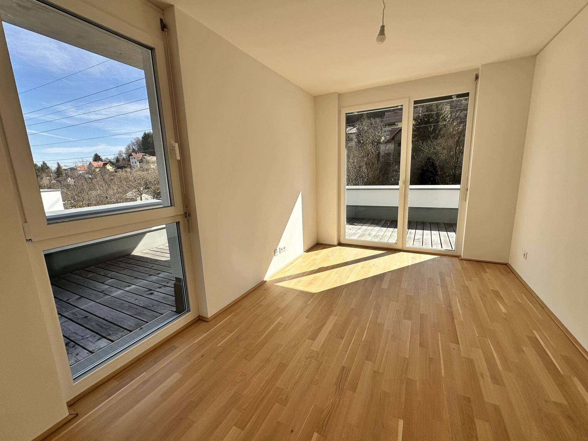Helles Zimmer mit Holzboden und großen Fenstern, die Zugang zu einer Außenterrasse bieten.