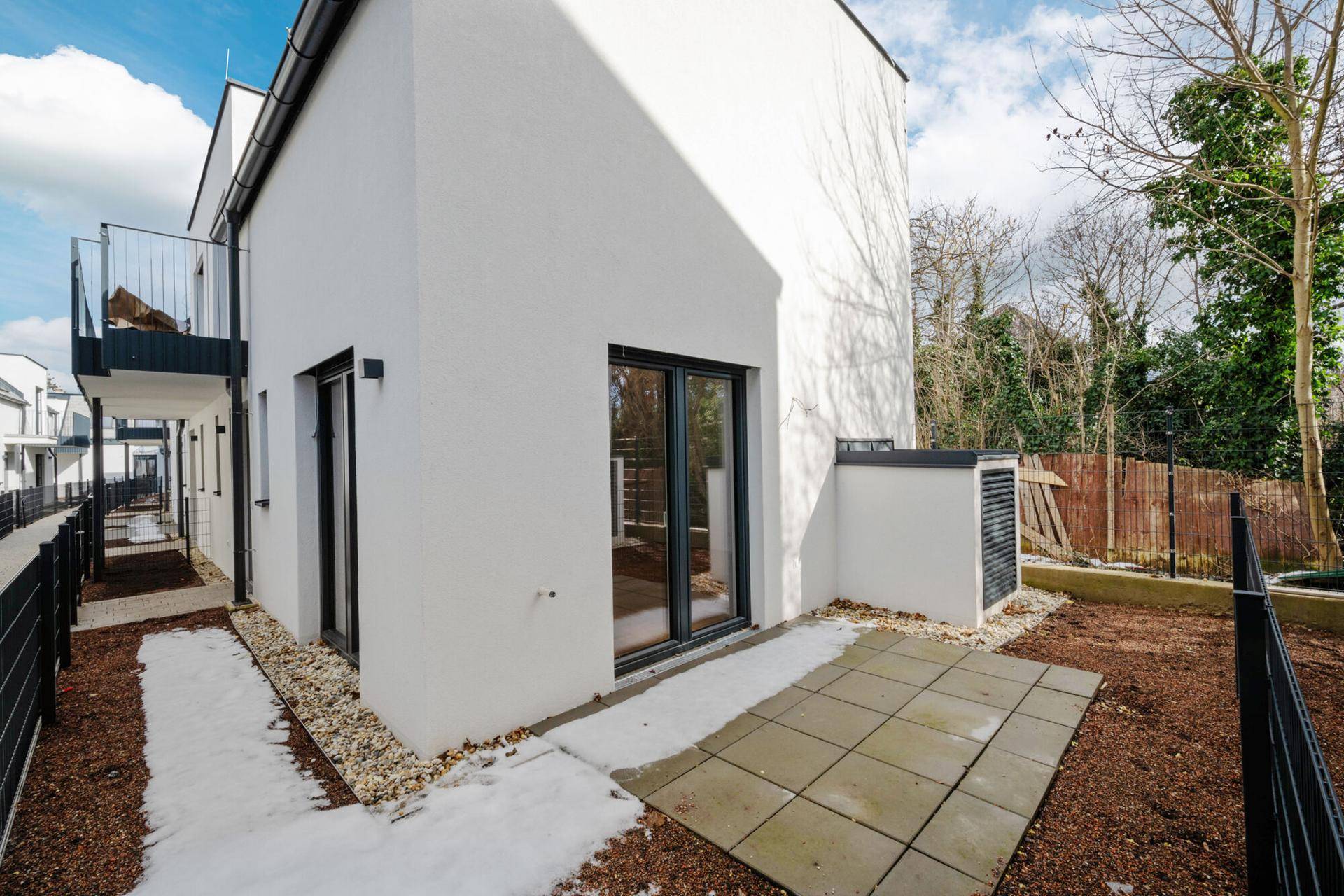 Seitliche Ansicht des Neubaus mit kleiner gepflasterter Terrasse und angrenzendem Gartenbereich.