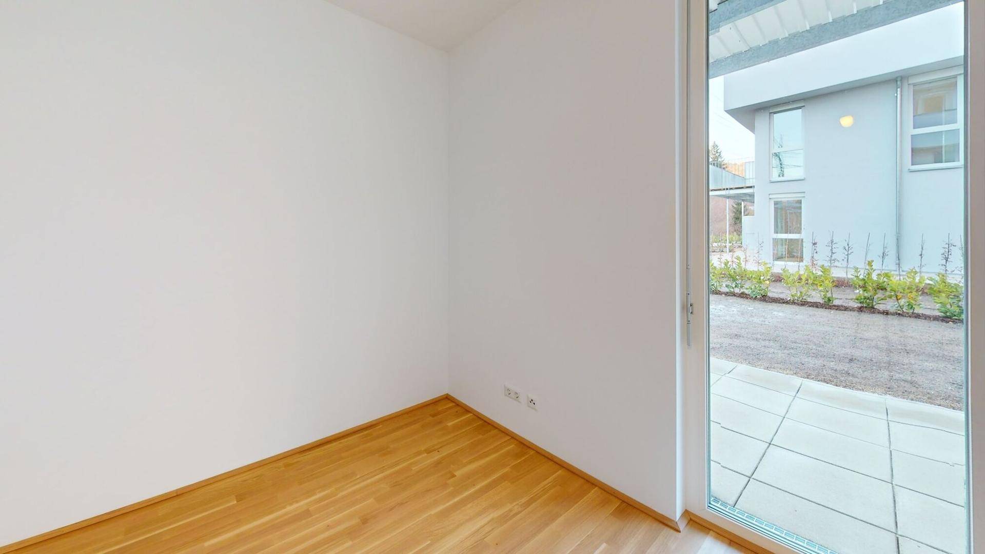 Unmöblierter Raum mit Holzboden und Zugang zu einer kleinen Terrasse mit Blick ins Grüne.