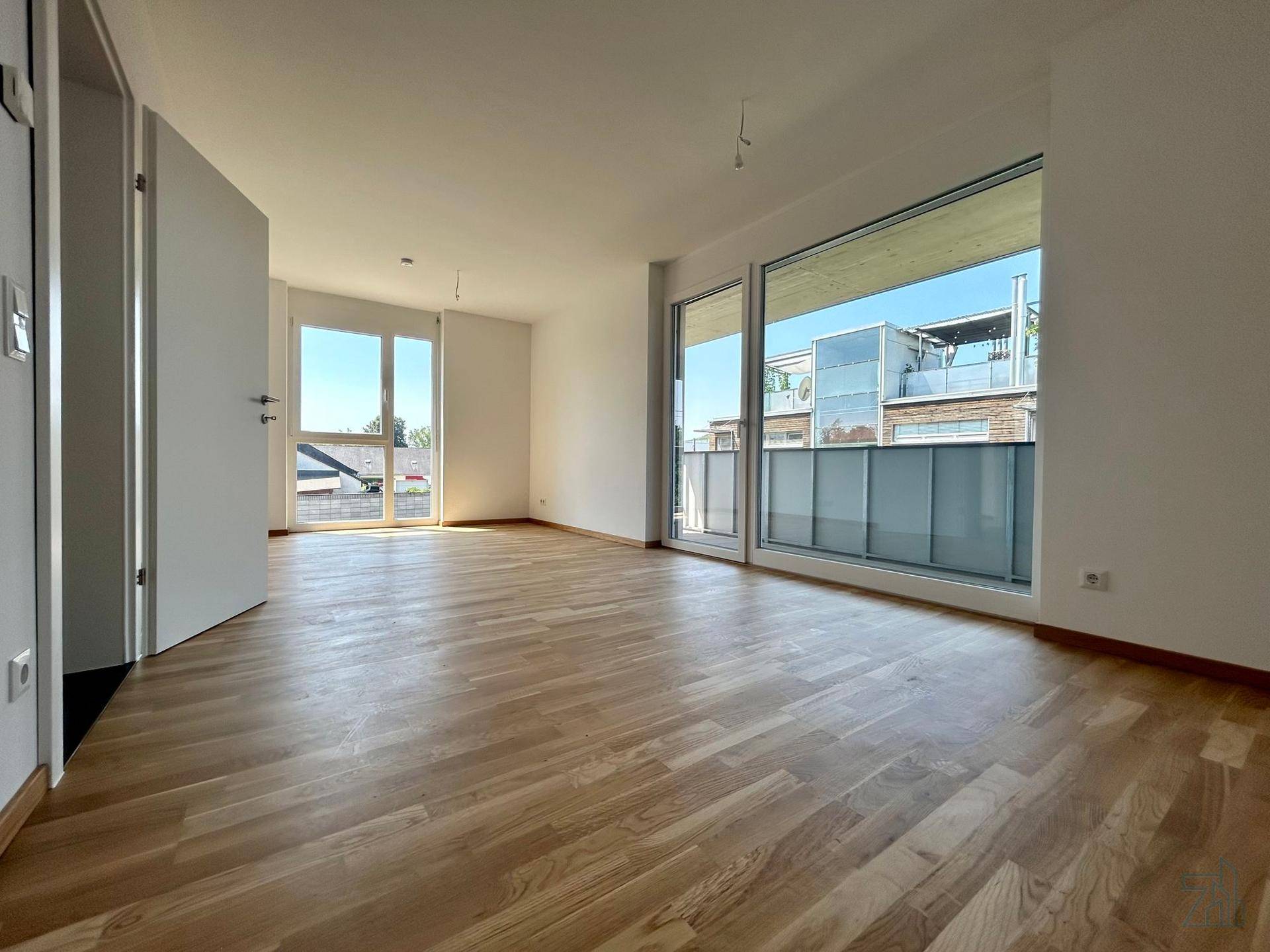 Geräumiger, heller Wohnraum mit großen Fenstern, Holzboden und viel Tageslicht.