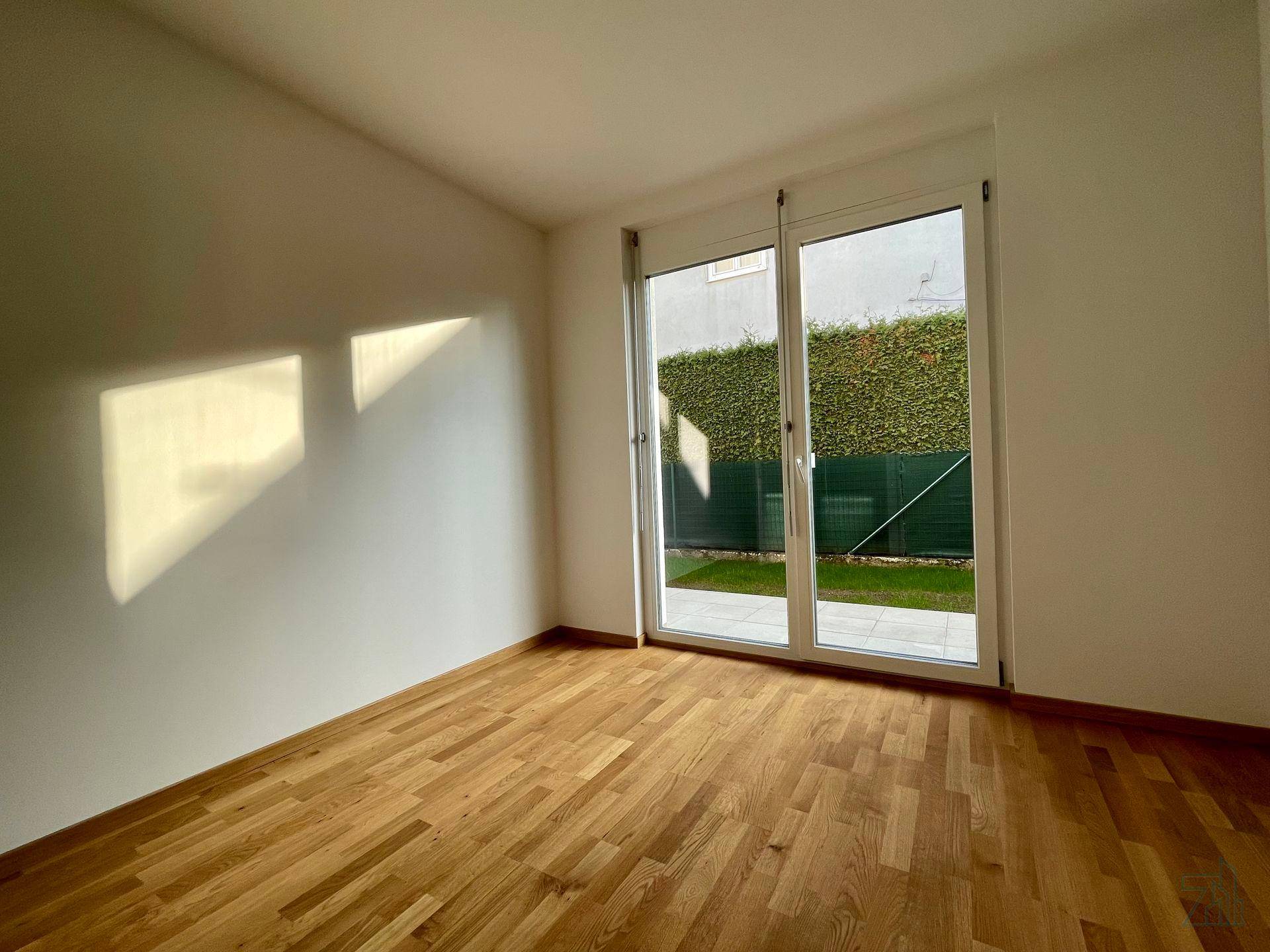 Leeres Zimmer mit Holzboden und großer Glastür, durch die Sonnenlicht auf die Wand fällt.
