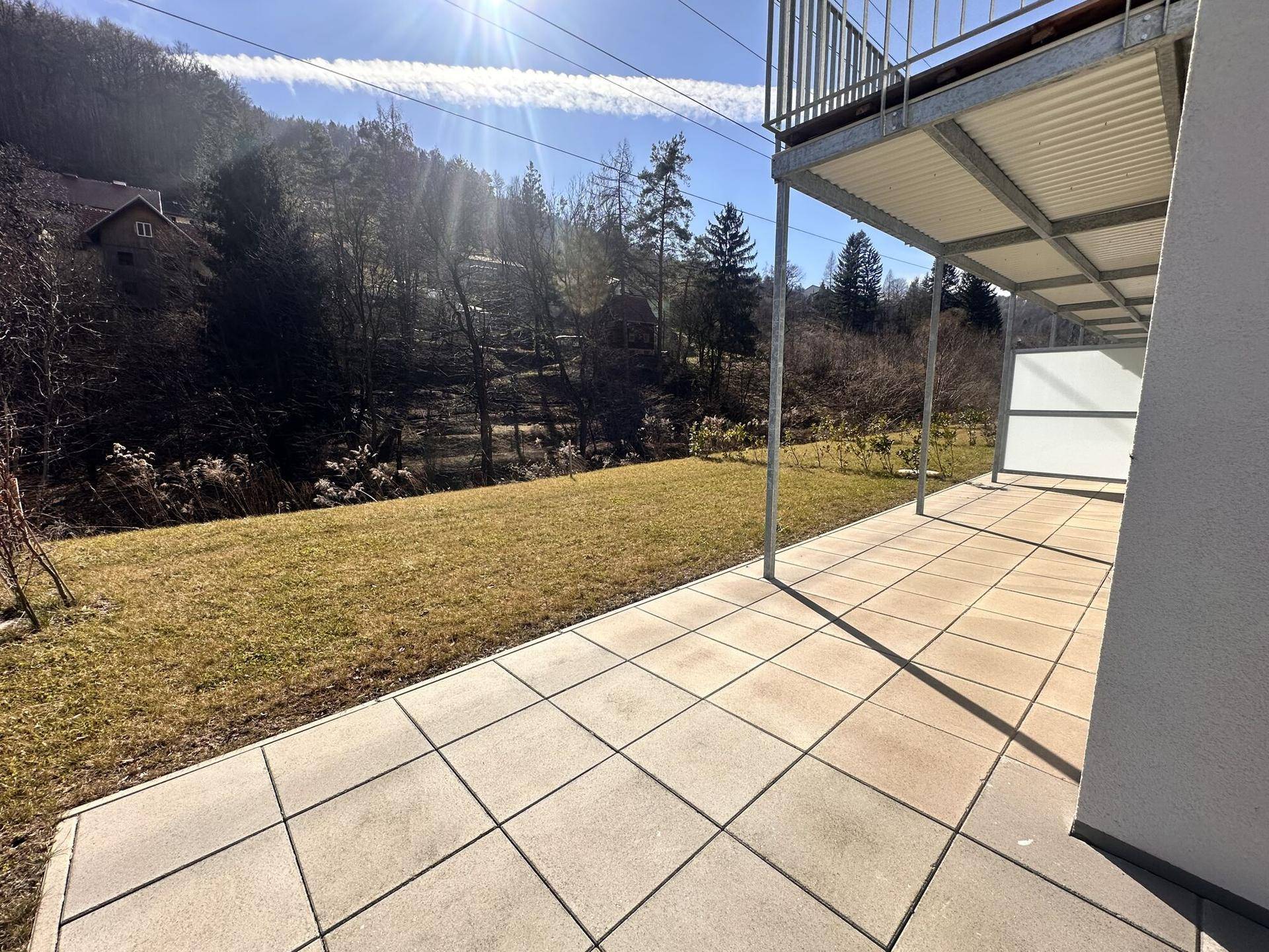 Sonnige Terrasse mit großen Pflastersteinen, angrenzender Rasenfläche und weitem Blick auf bewaldete Hügel.
