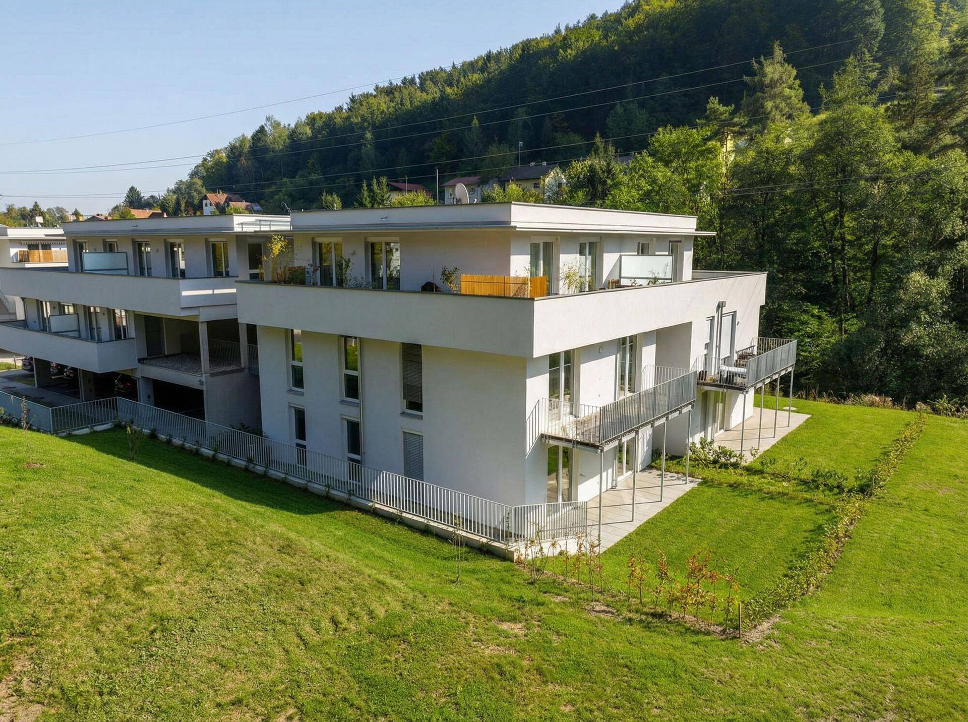 Luftaufnahme einer modernen Wohnanlage mit Flachdächern und umliegender Natur vor bewaldeten Bergen.