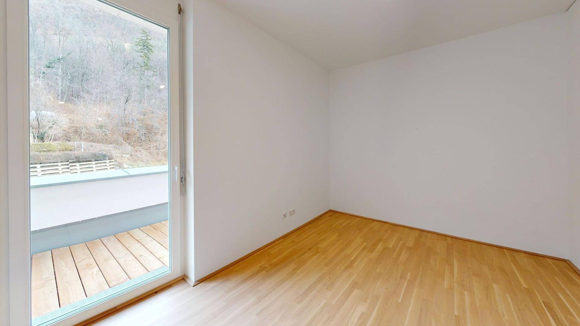 Leeres Zimmer mit hellem Parkettboden und Zugang zu einem Balkon mit Blick ins Grüne.