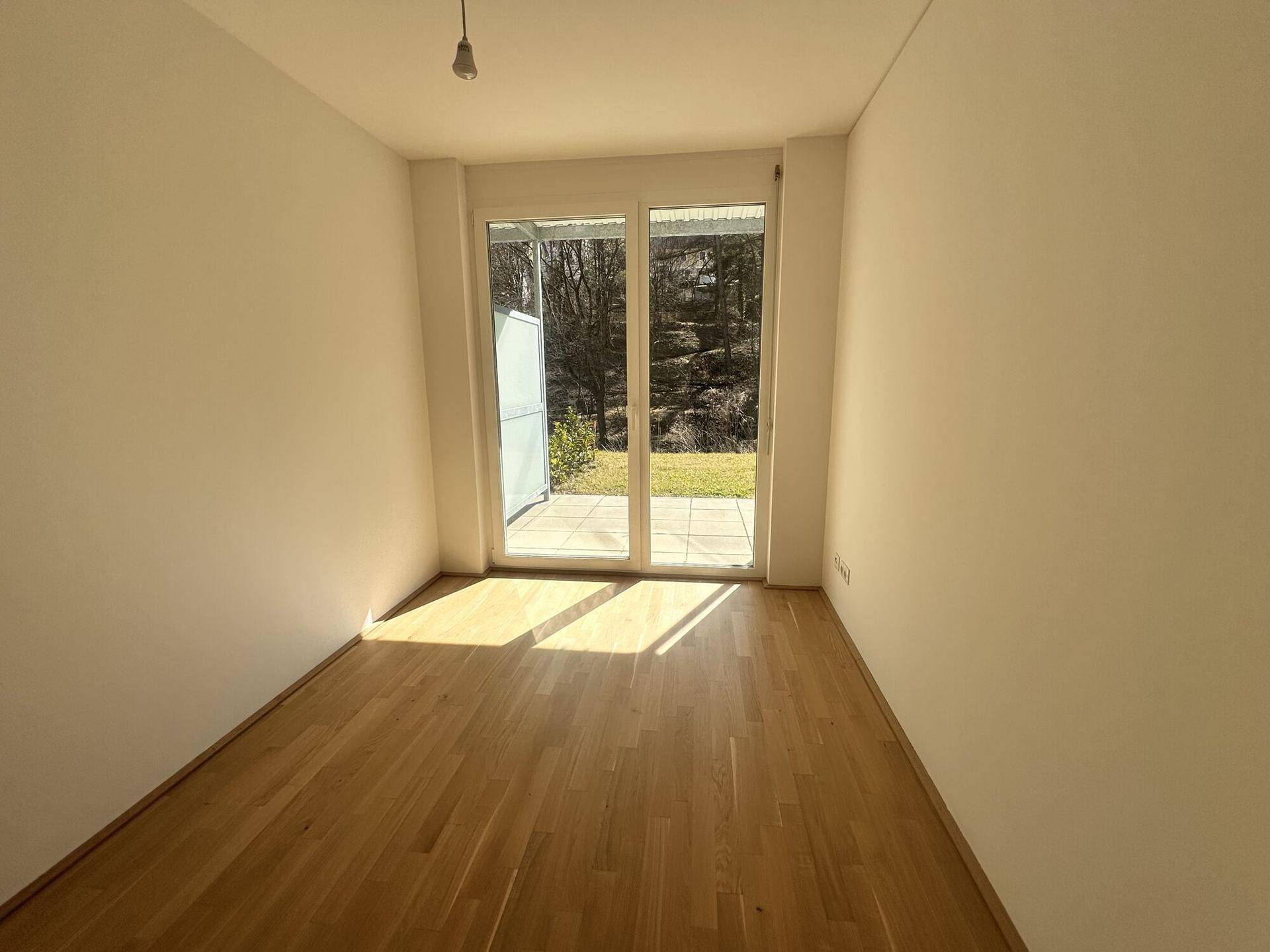 Heller Raum mit Holzboden und großer Terrassentür, die viel Licht hereinlässt.