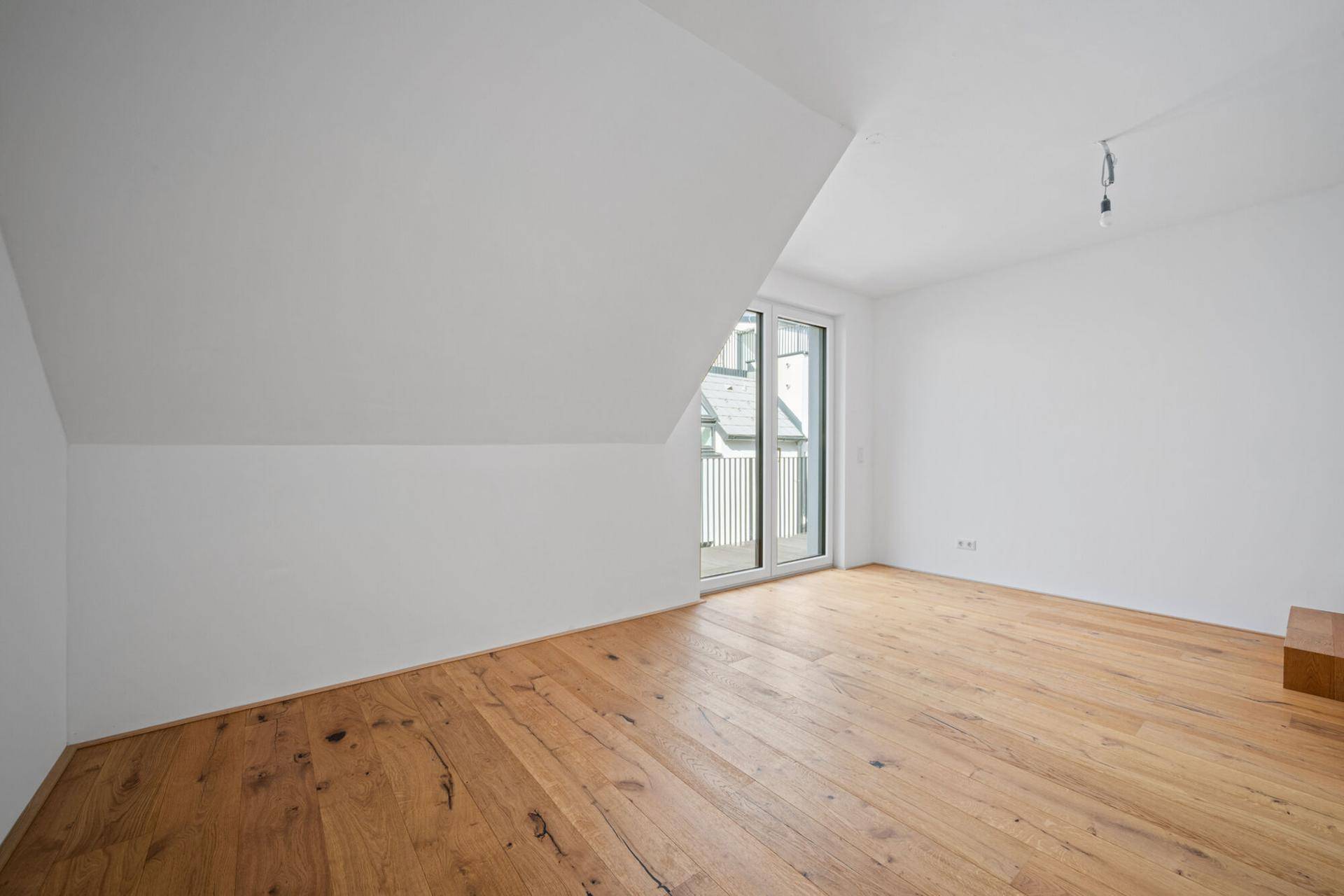 Helles Zimmer mit Holzboden, Dachschräge und großem Fenster mit Zugang zum Balkon.