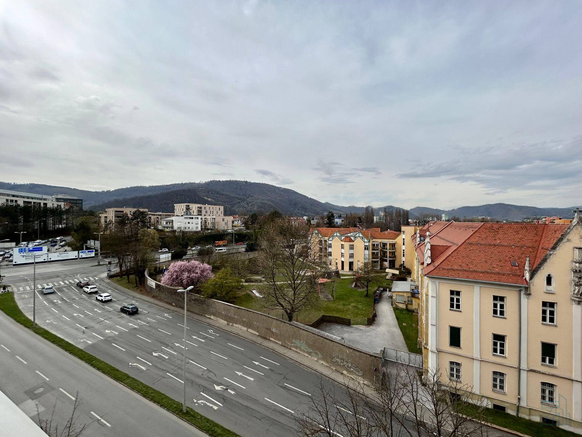 Weitläufiger Blick über eine Stadtstraße mit Gebäuden und grünen Hügeln im Hintergrund.