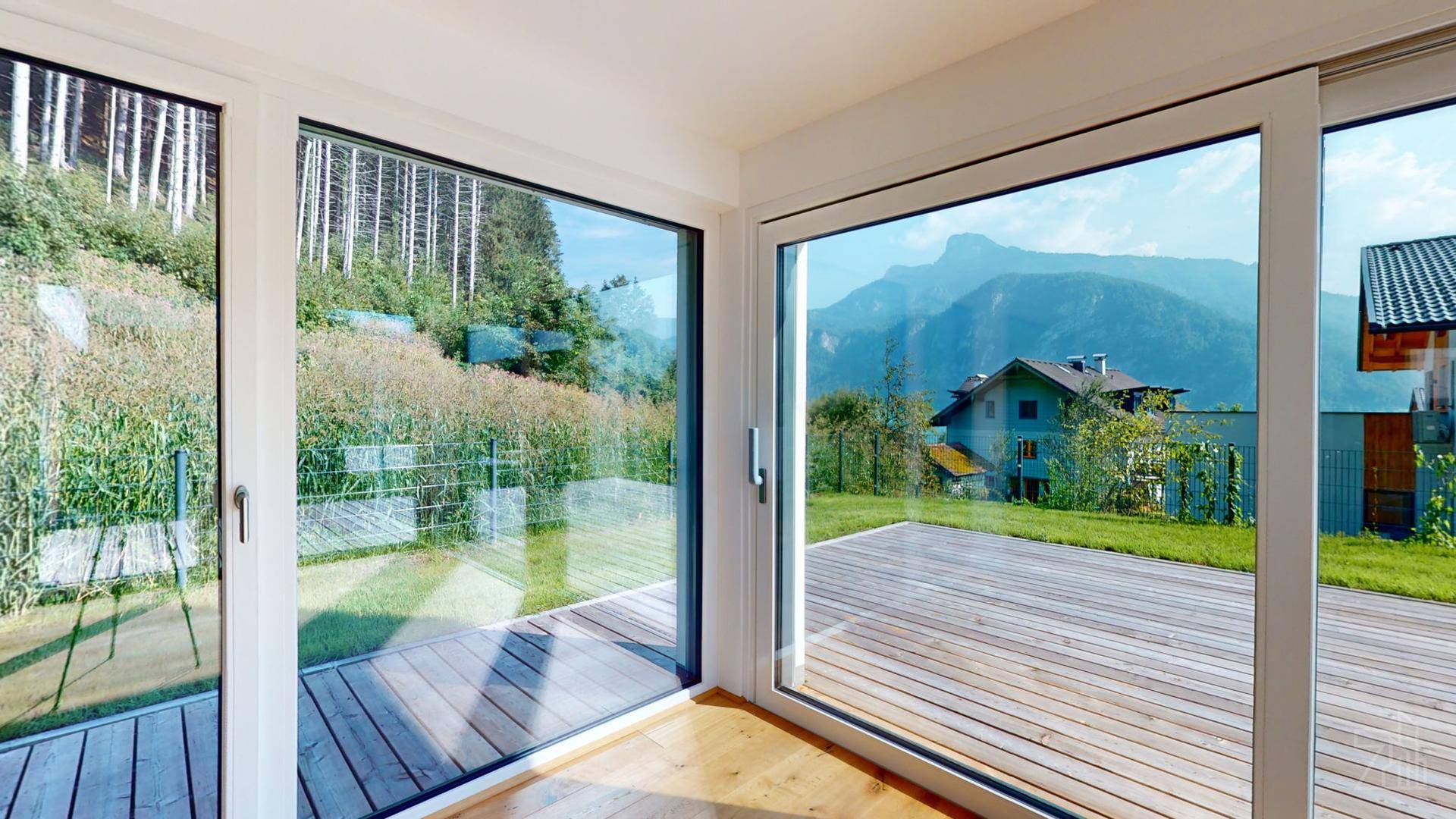 Heller Innenraum mit großen Fenstern, die einen weiten Blick auf die Berge und die Holzterrasse bieten.